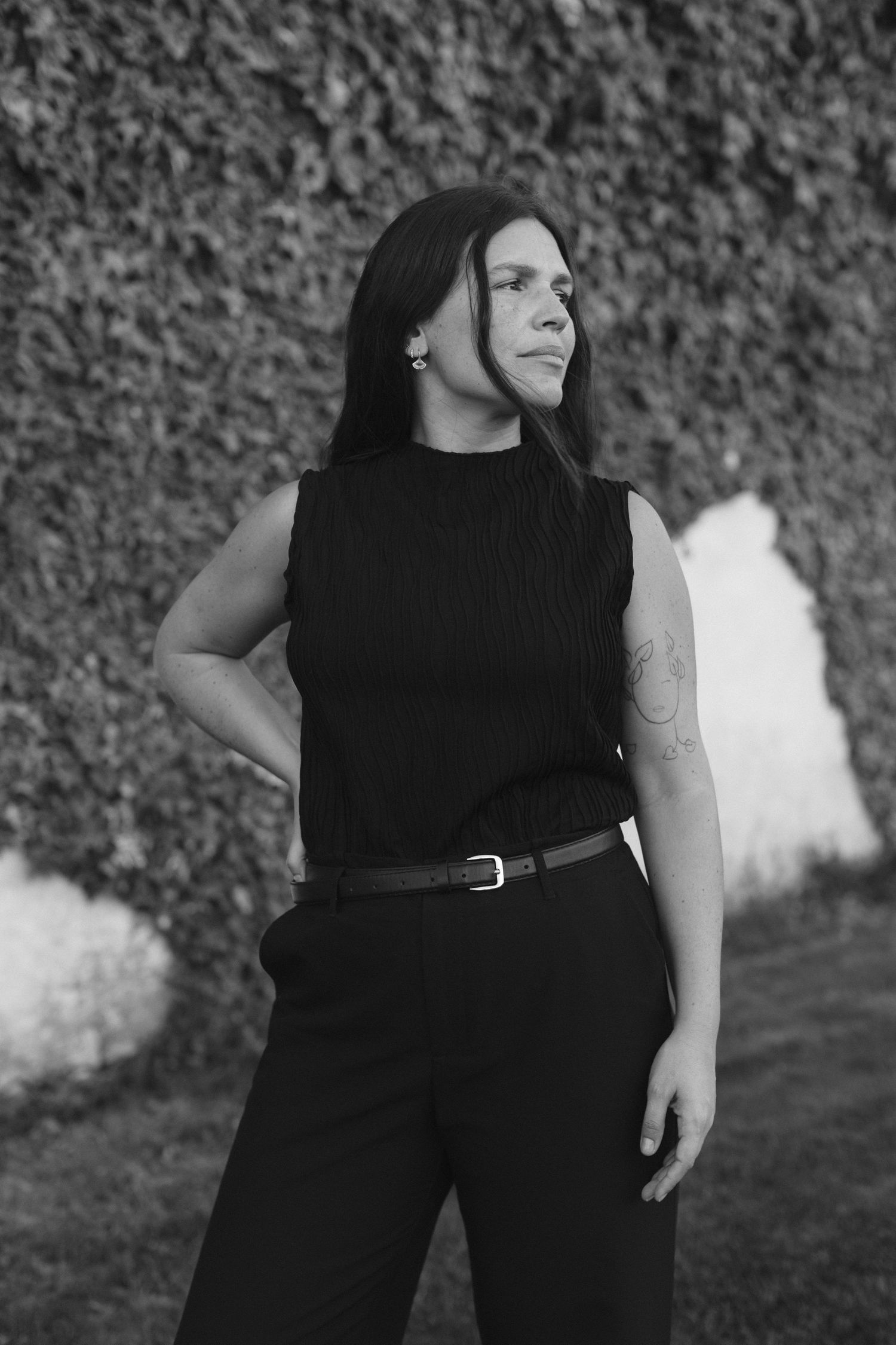 Black and white portrait of a woman in black sleeveless outfit with belt, standing outdoors against ivy wall.