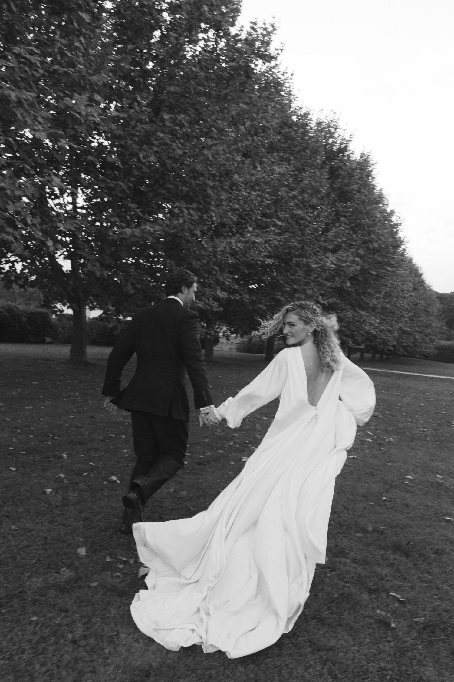 A flowing white dress billows dramatically in the wind across a grassy field with trees in the background.