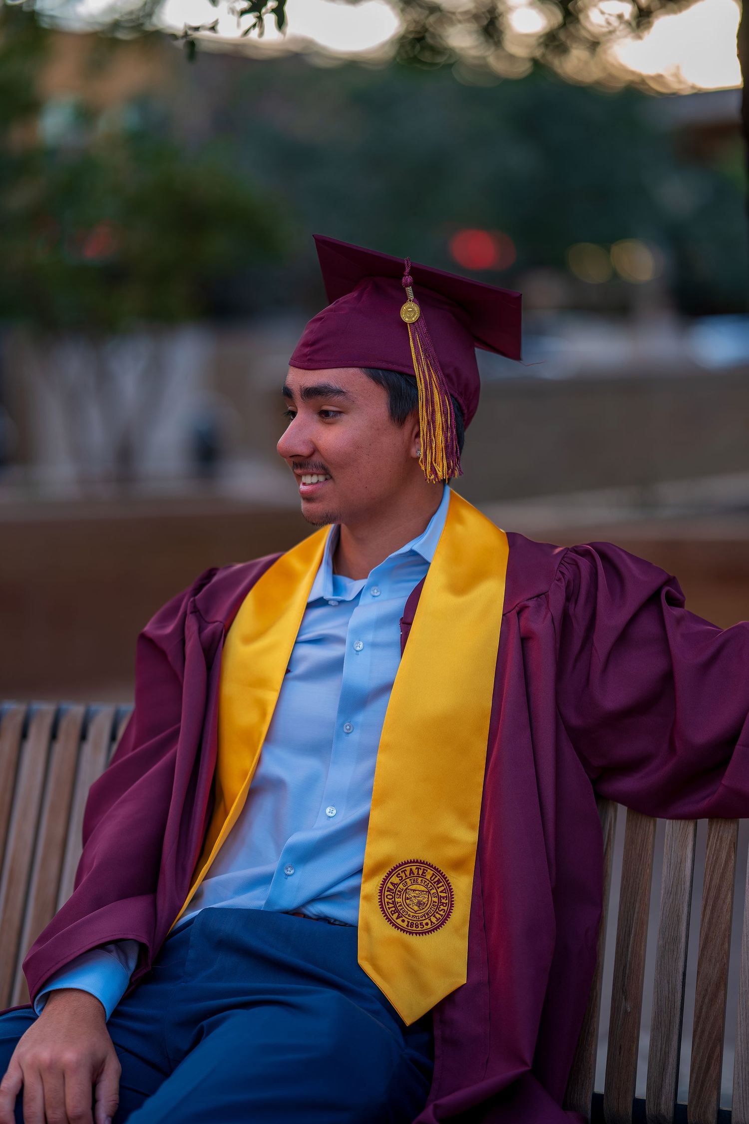 Graduate in maroon and gold regalia relaxing on outdoor bench.