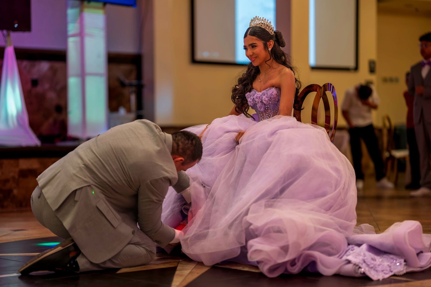 A quinceañera ceremony with a girl in a lavender ball gown while a man kneels before her on a dance floor.