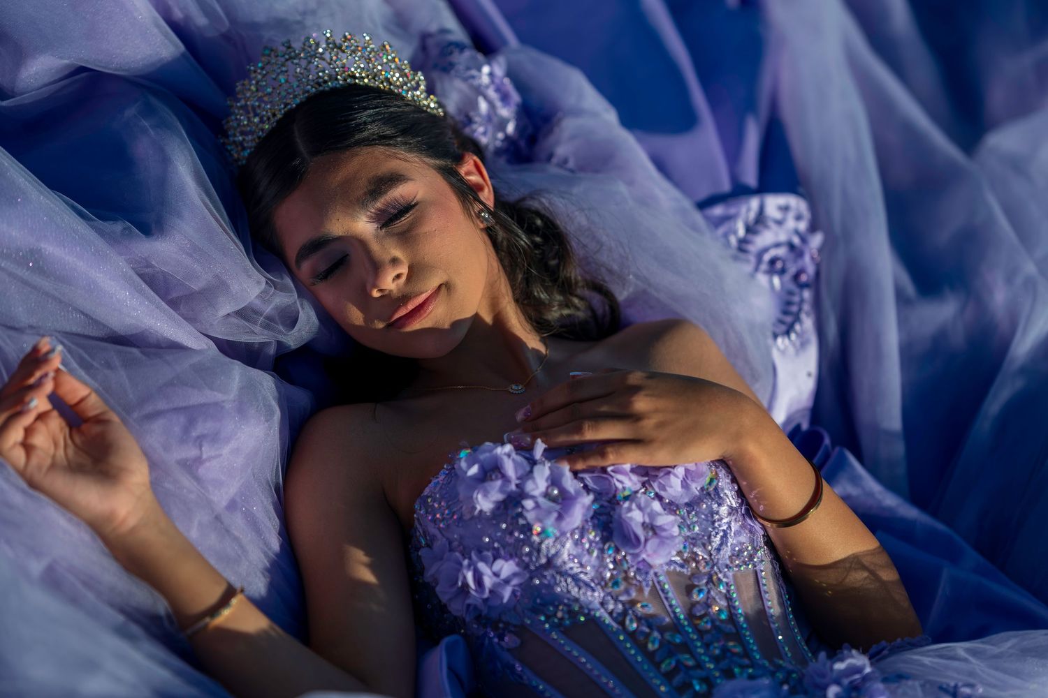 Young woman in lavender quinceañera gown and tiara lying gracefully among flowing purple fabric.