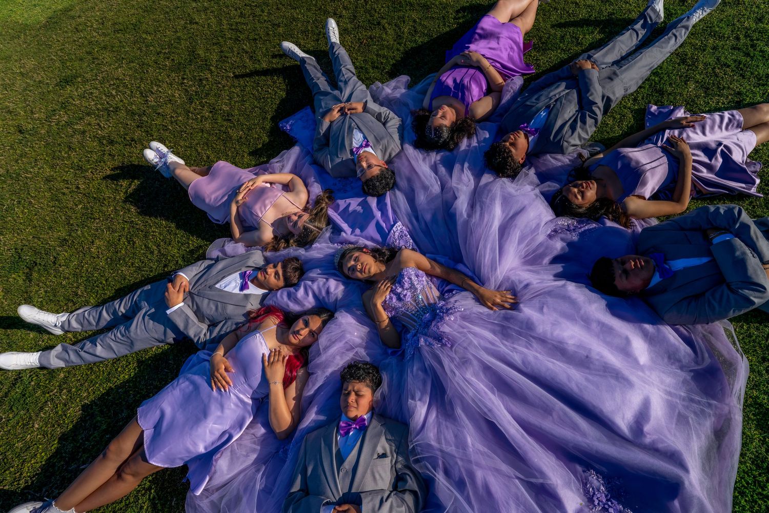 Overhead view of quinceañera court lying on grass around girl in lavender ball gown celebration.