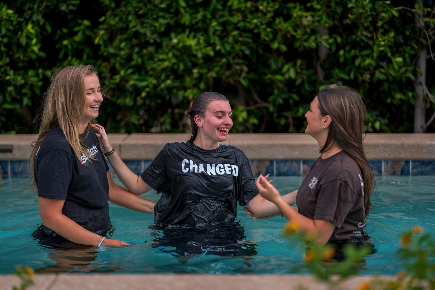 Three women stand in a pool, one wearing a 'CHANGED' shirt, sharing a joyful baptism moment.