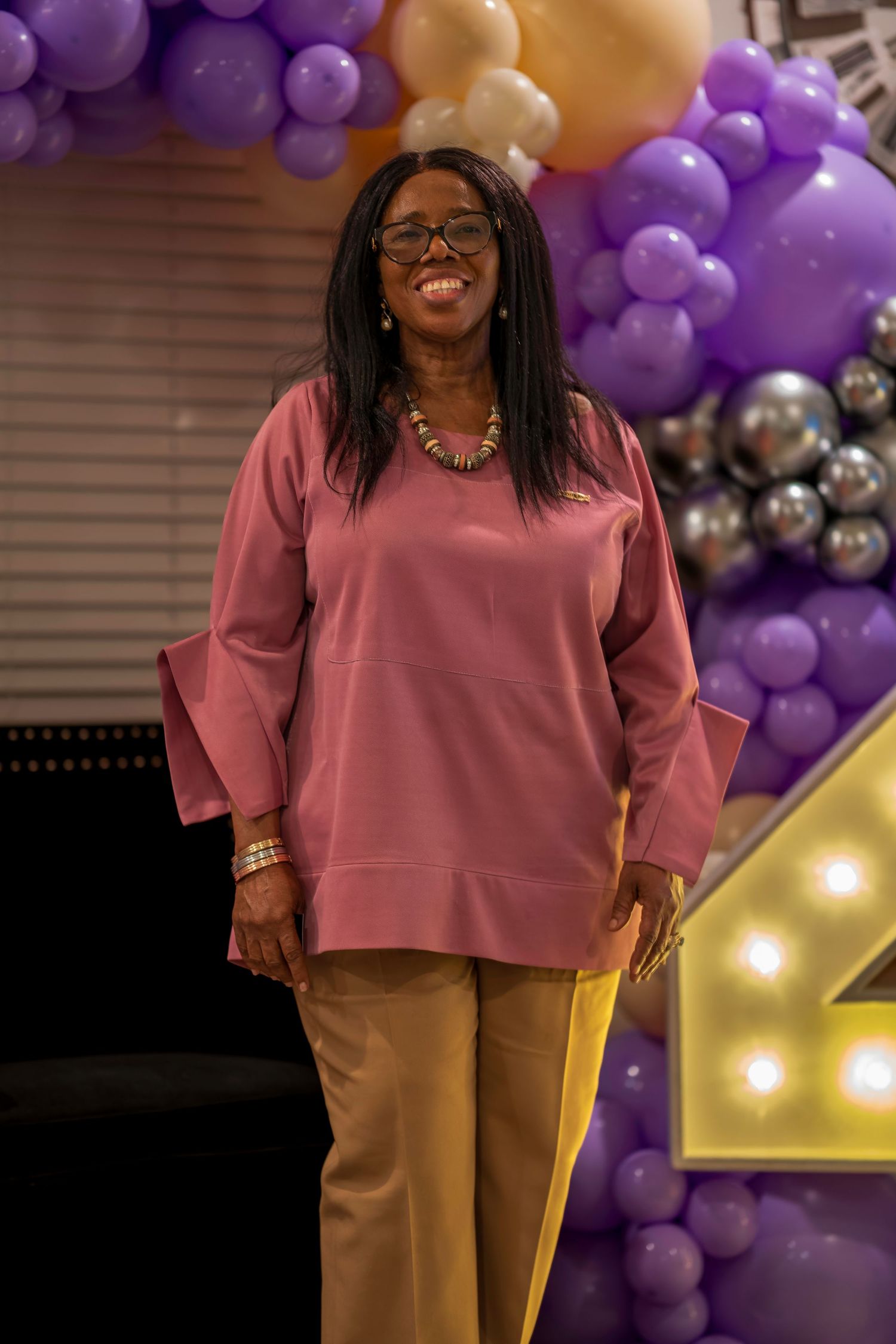 Smiling woman in pink top and yellow pants poses before purple and gold balloon arch.