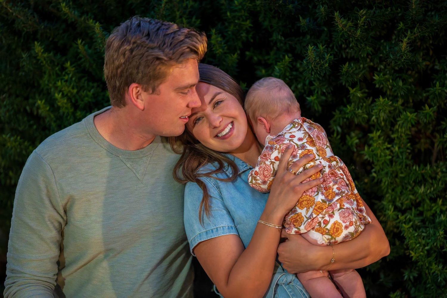 Happy couple sharing a tender moment while holding their newborn baby outdoors near green trees.