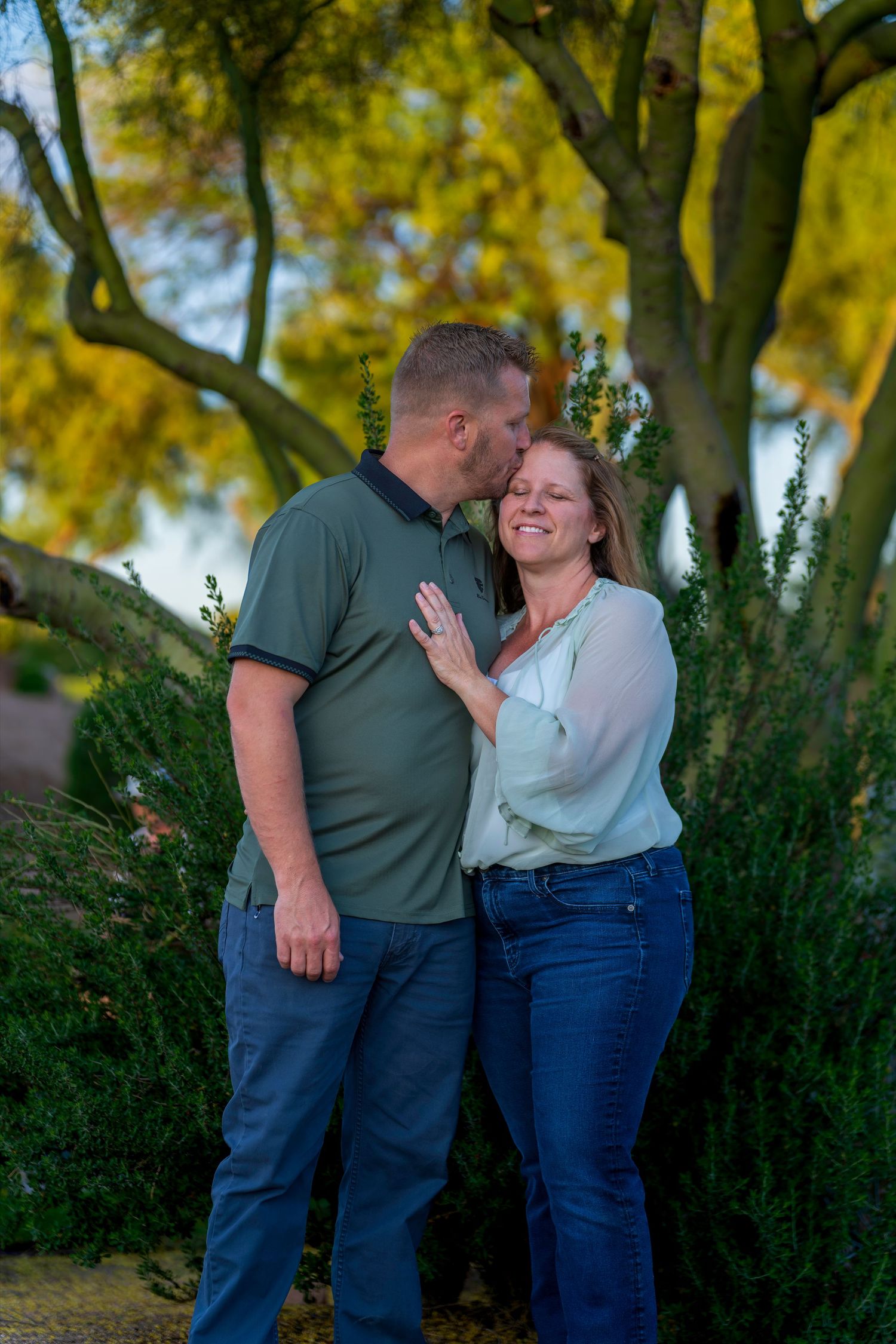 A couple shares a tender kiss outdoors, surrounded by lush green bushes and golden trees in warm evening light.