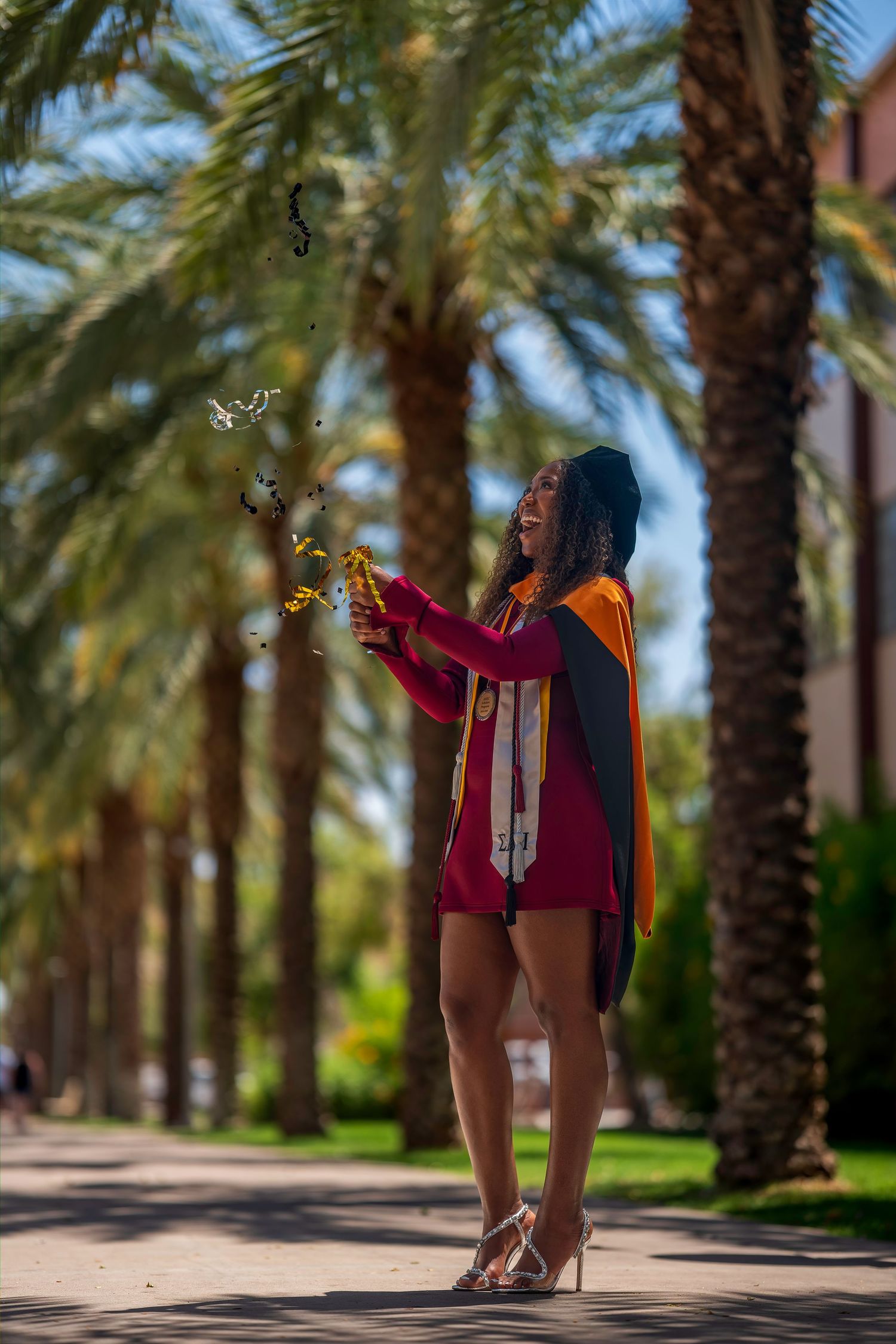 Joyful ASU graduate in maroon regalia popping confetti among palm trees on sunny campus walkway.