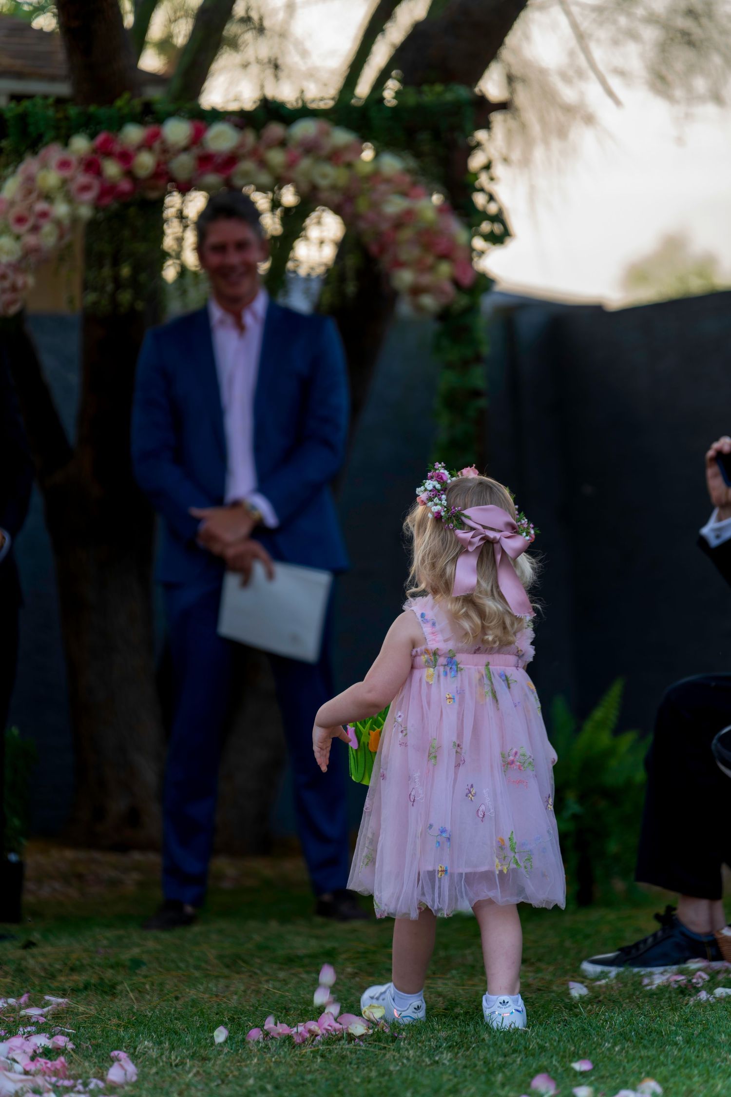 Flower girl in pink dress scattering petals at outdoor wedding ceremony with floral arch.