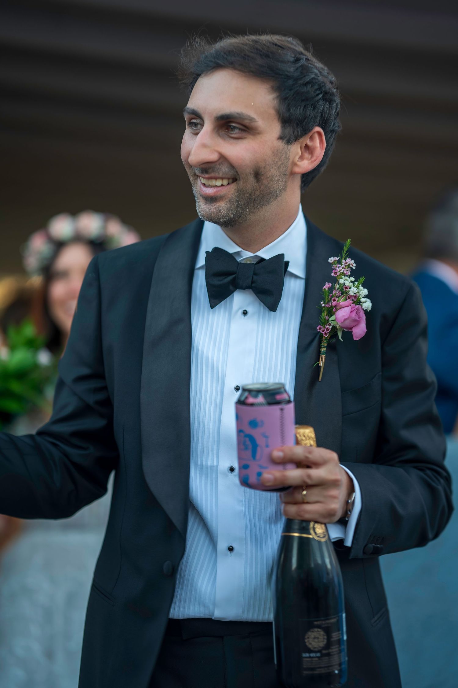 Smiling groom in black tuxedo with bow tie and floral boutonniere holding champagne bottle and beer can at wedding.