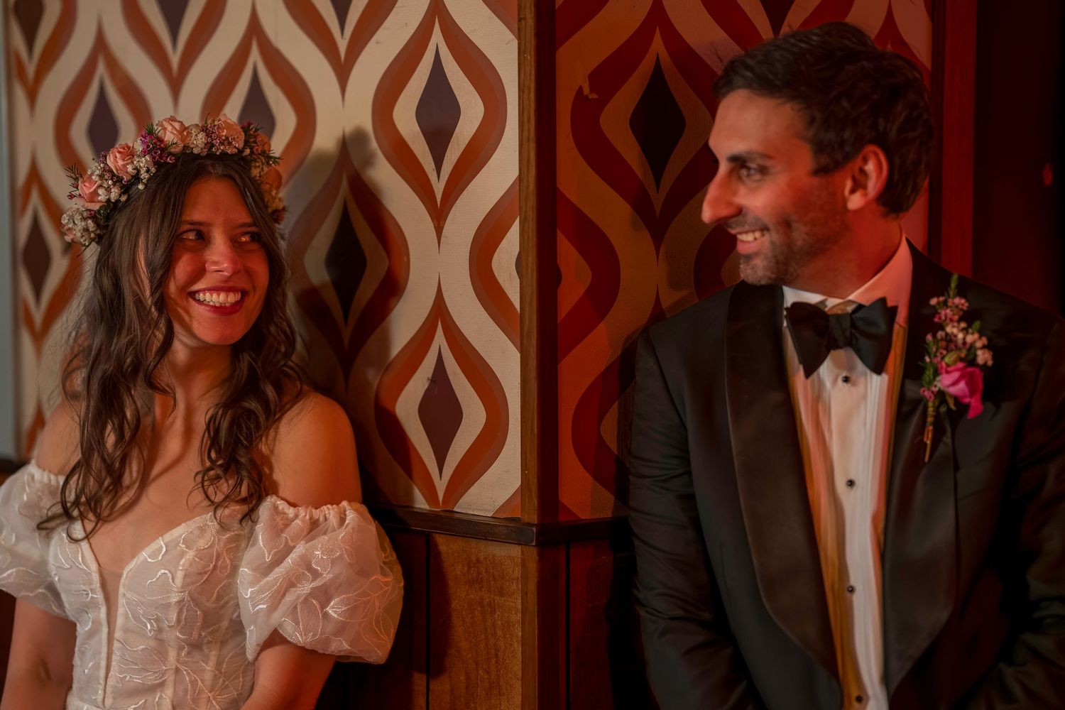 Bride in floral crown and white gown smiles beside groom in black tuxedo with boutonniere at wedding.