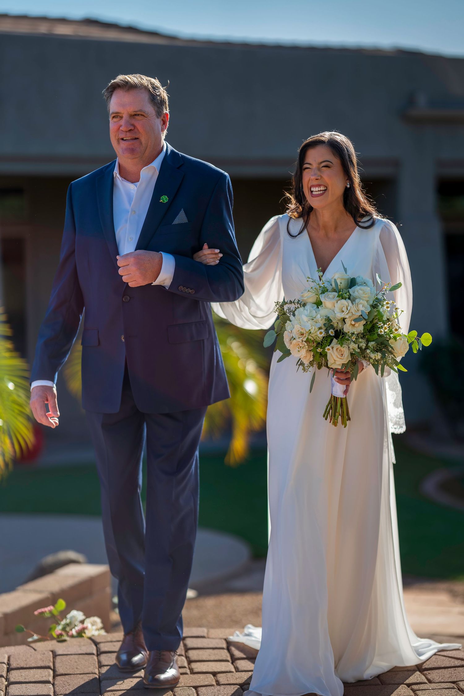 Smiling bride in white dress holding bouquet walks arm-in-arm with man in blue suit outdoors.