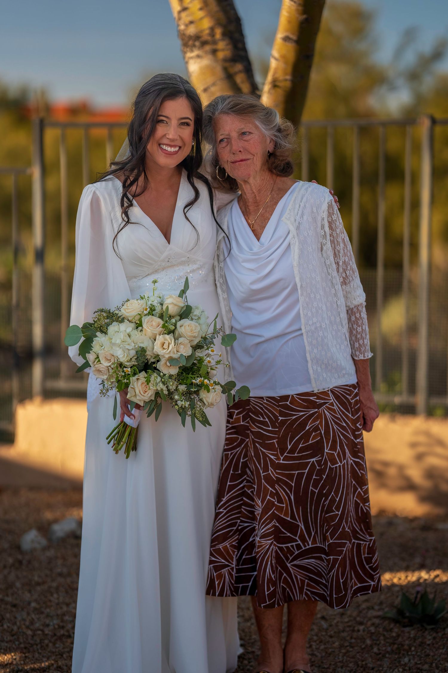 Radiant bride in white gown holding white bouquet poses with elderly woman in warm golden outdoor light.
