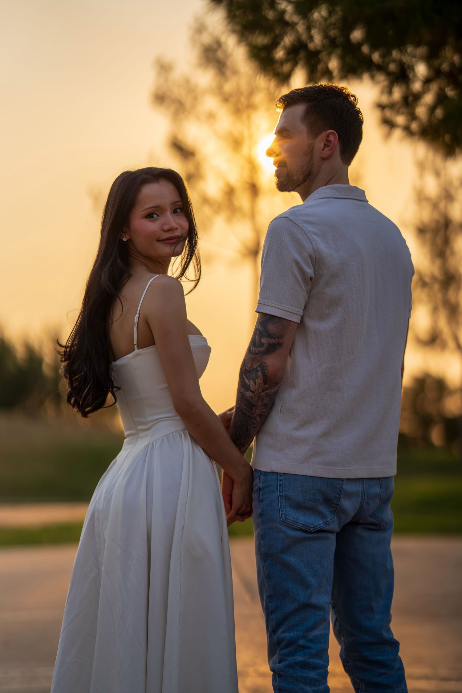 Couple holding hands at golden hour sunset, woman in white dress smiling at camera.