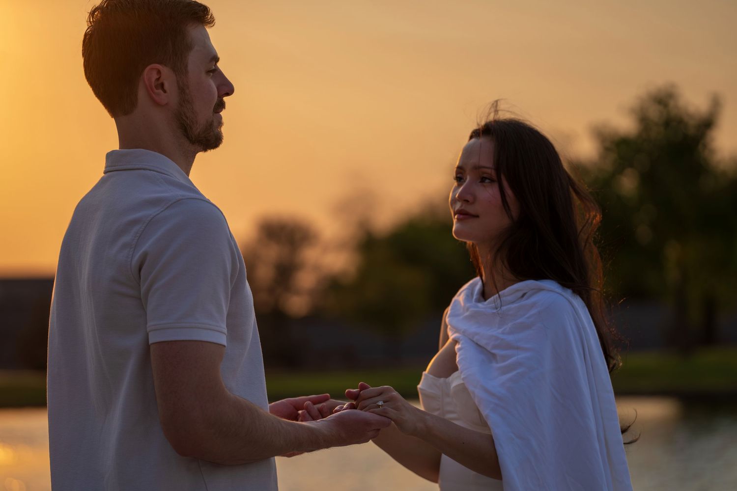 Couple holding hands during a golden sunset proposal moment near a serene waterfront.