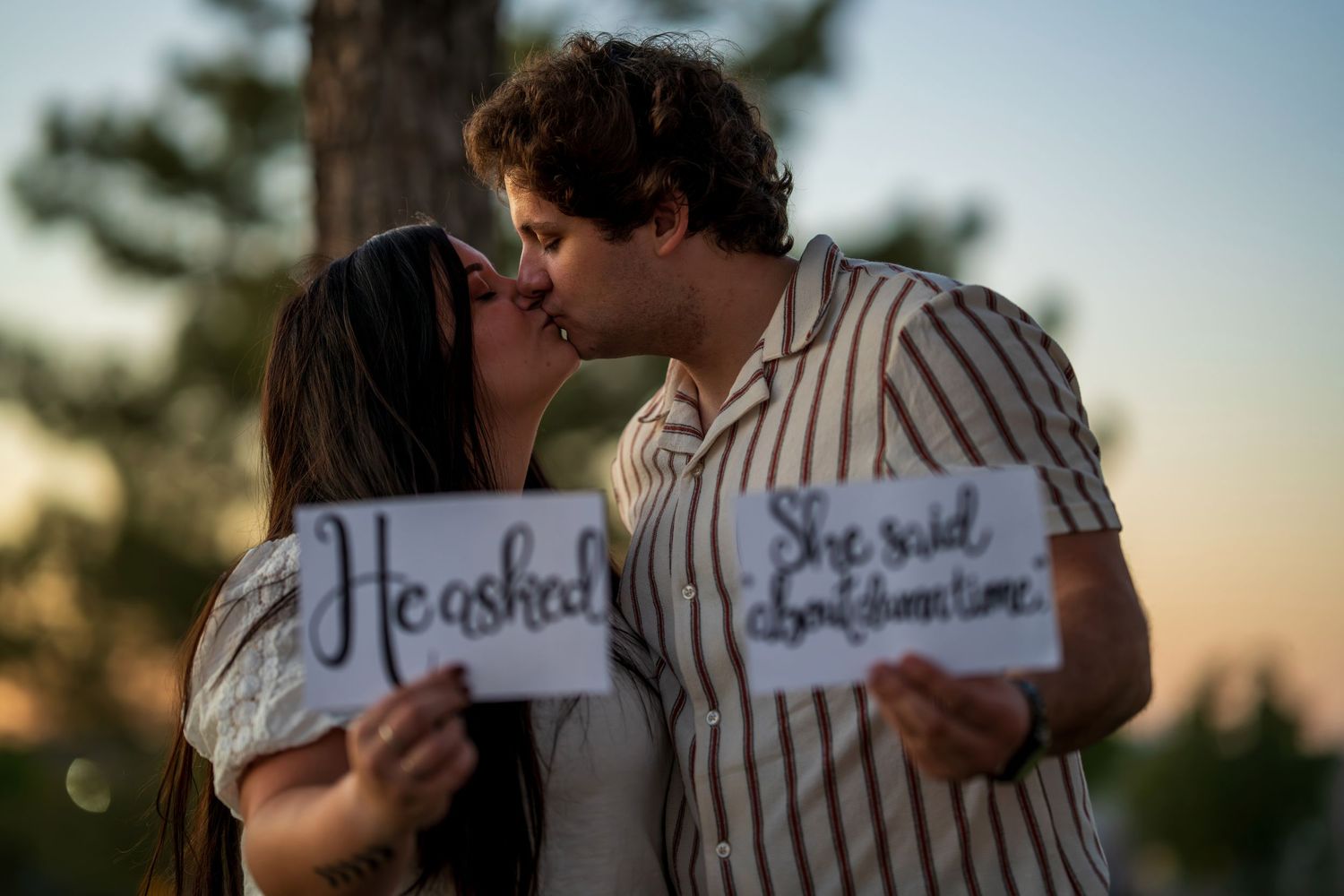 Couple kissing at sunset holding handwritten signs announcing their engagement outdoors.