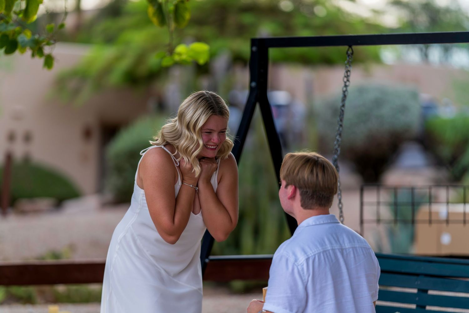 Emotional woman in white dress covering her mouth in surprise during an outdoor marriage proposal on a swing.