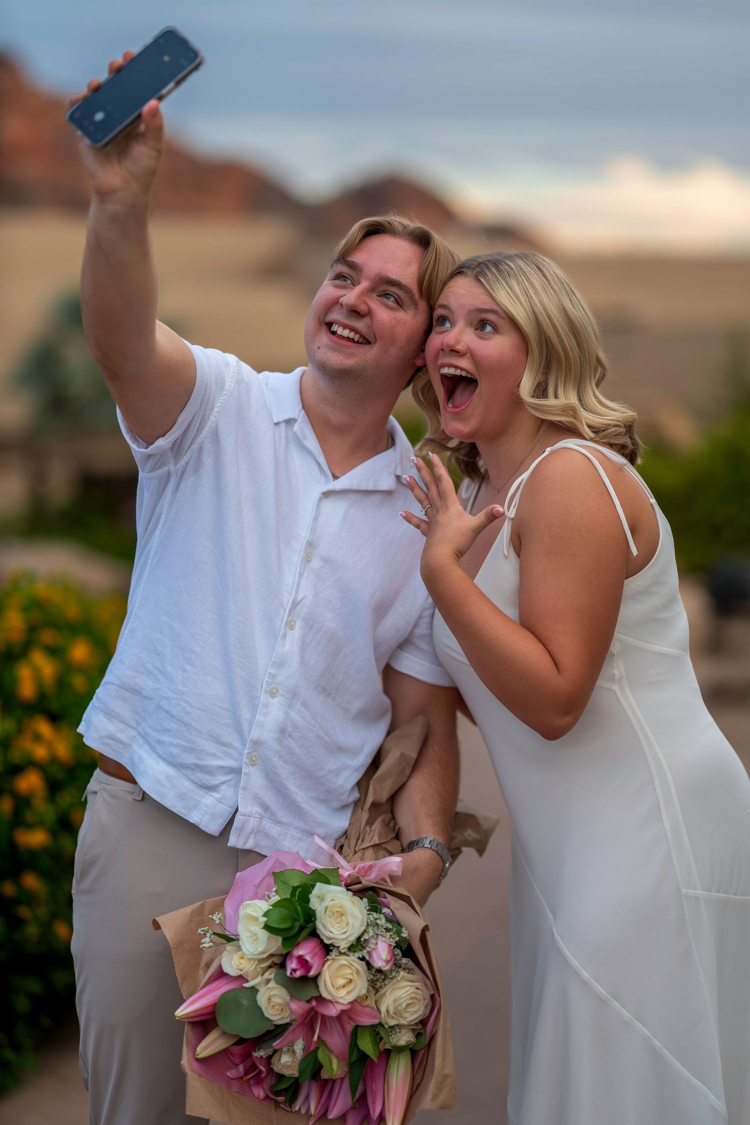 Excited couple in white taking a selfie outdoors, woman holding a bouquet of pink and white roses.