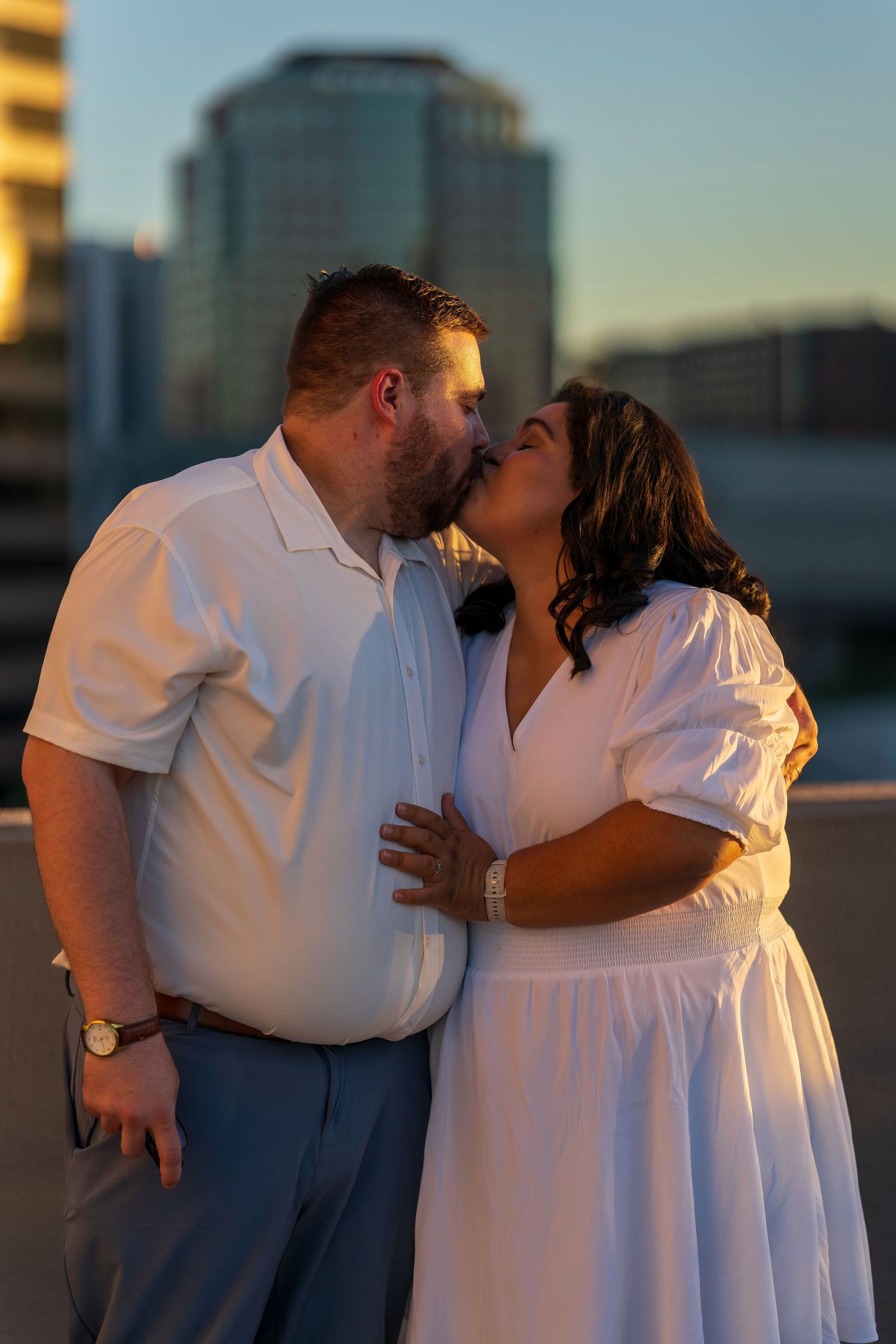 Couple sharing a romantic kiss on a rooftop at sunset, wearing white outfits with city buildings in the background.