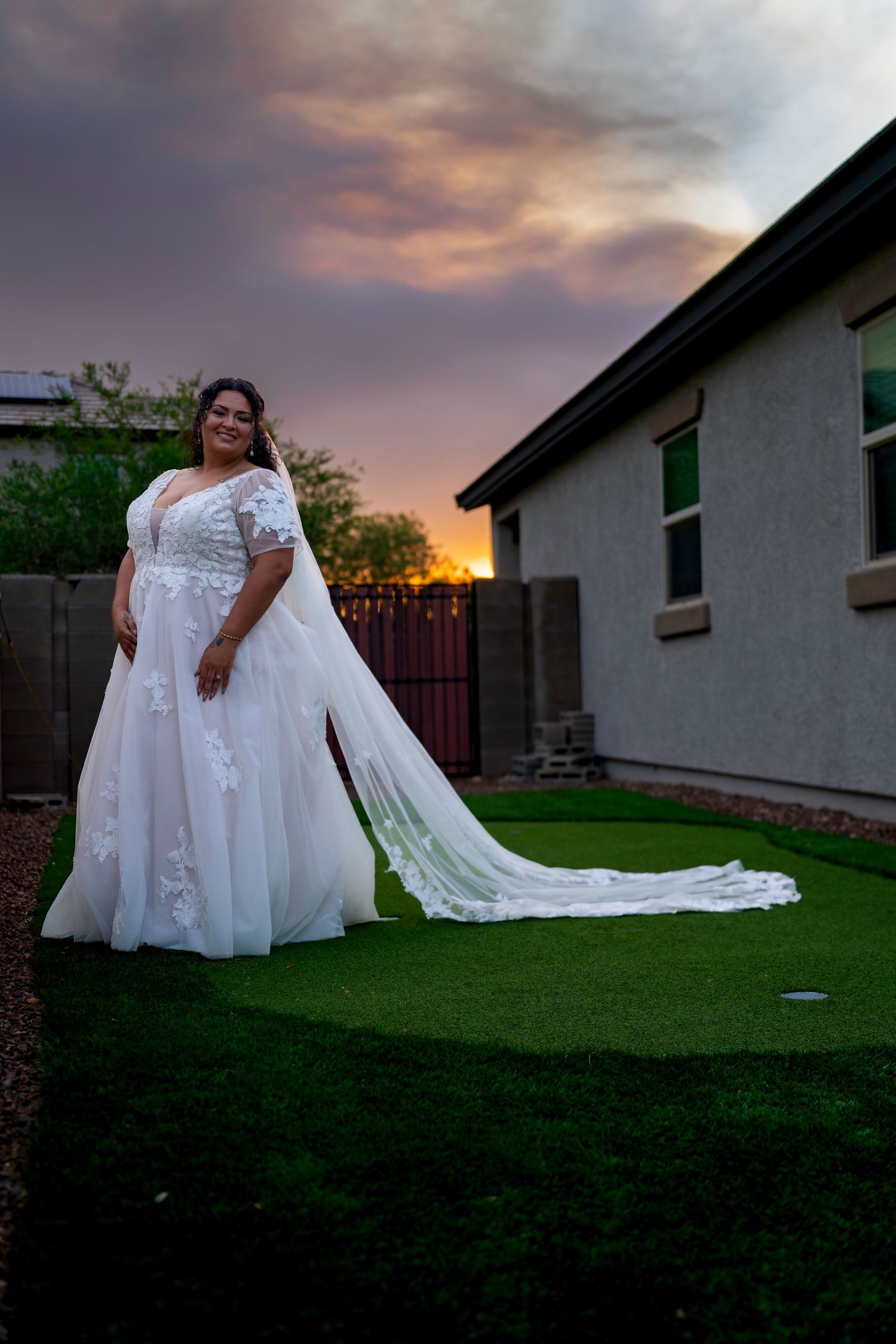Bride in elegant white lace gown with long train poses solo on green lawn at dramatic sunset wedding.