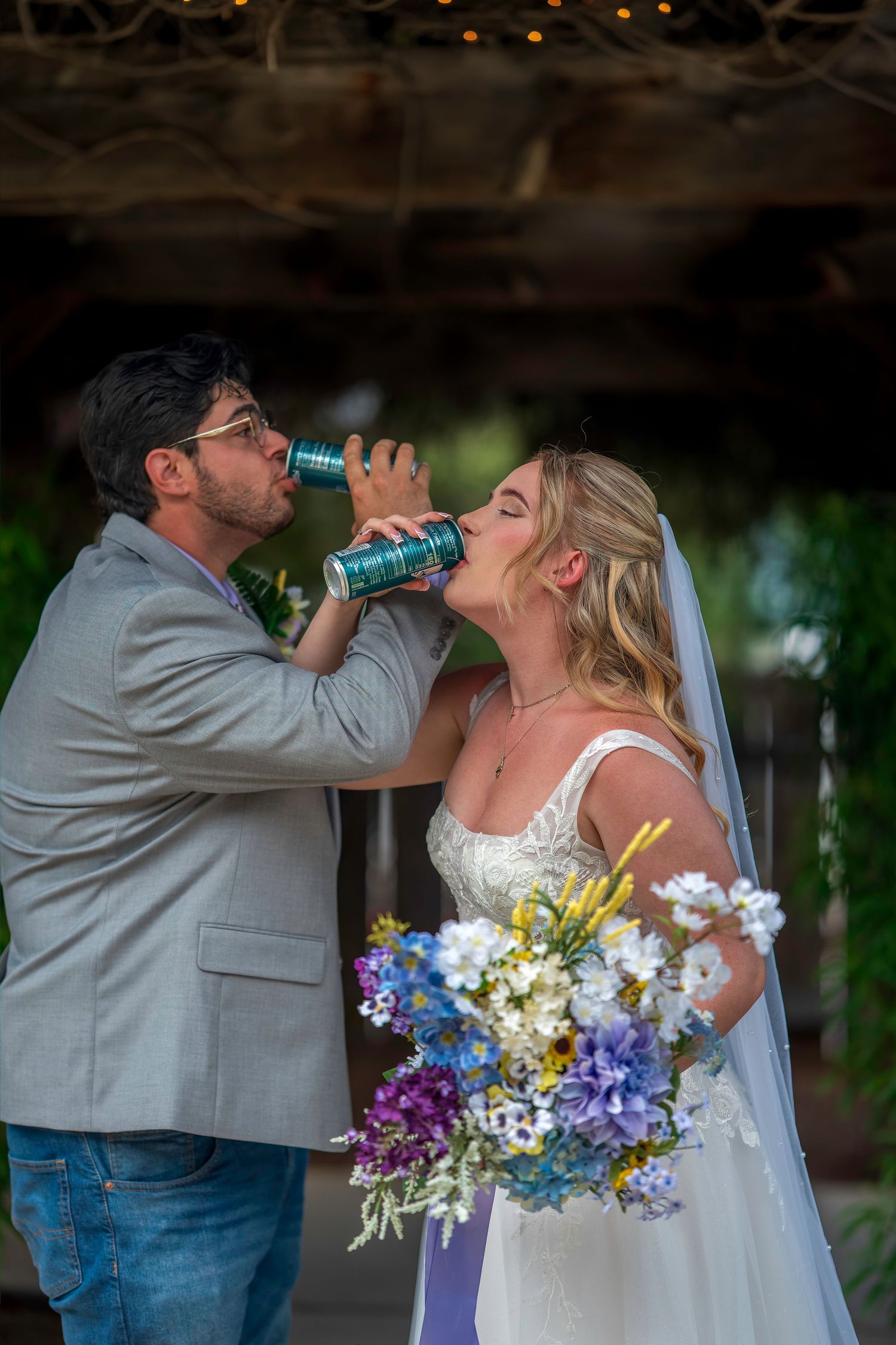 Bride and groom sharing a drink from a can during their outdoor wedding ceremony with floral bouquet.