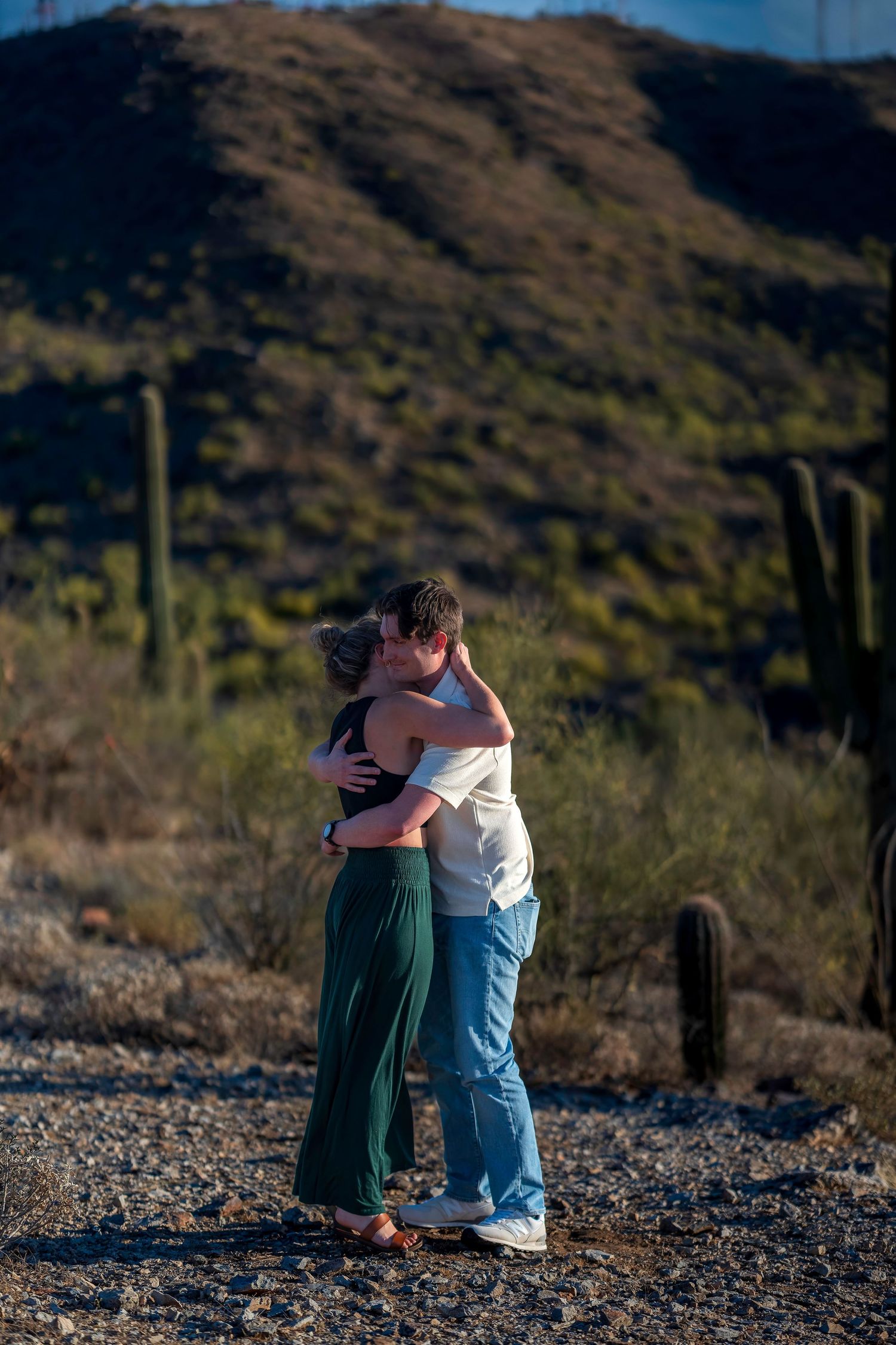 Couple embracing and kissing in a desert landscape with saguaro cacti and mountain backdrop.