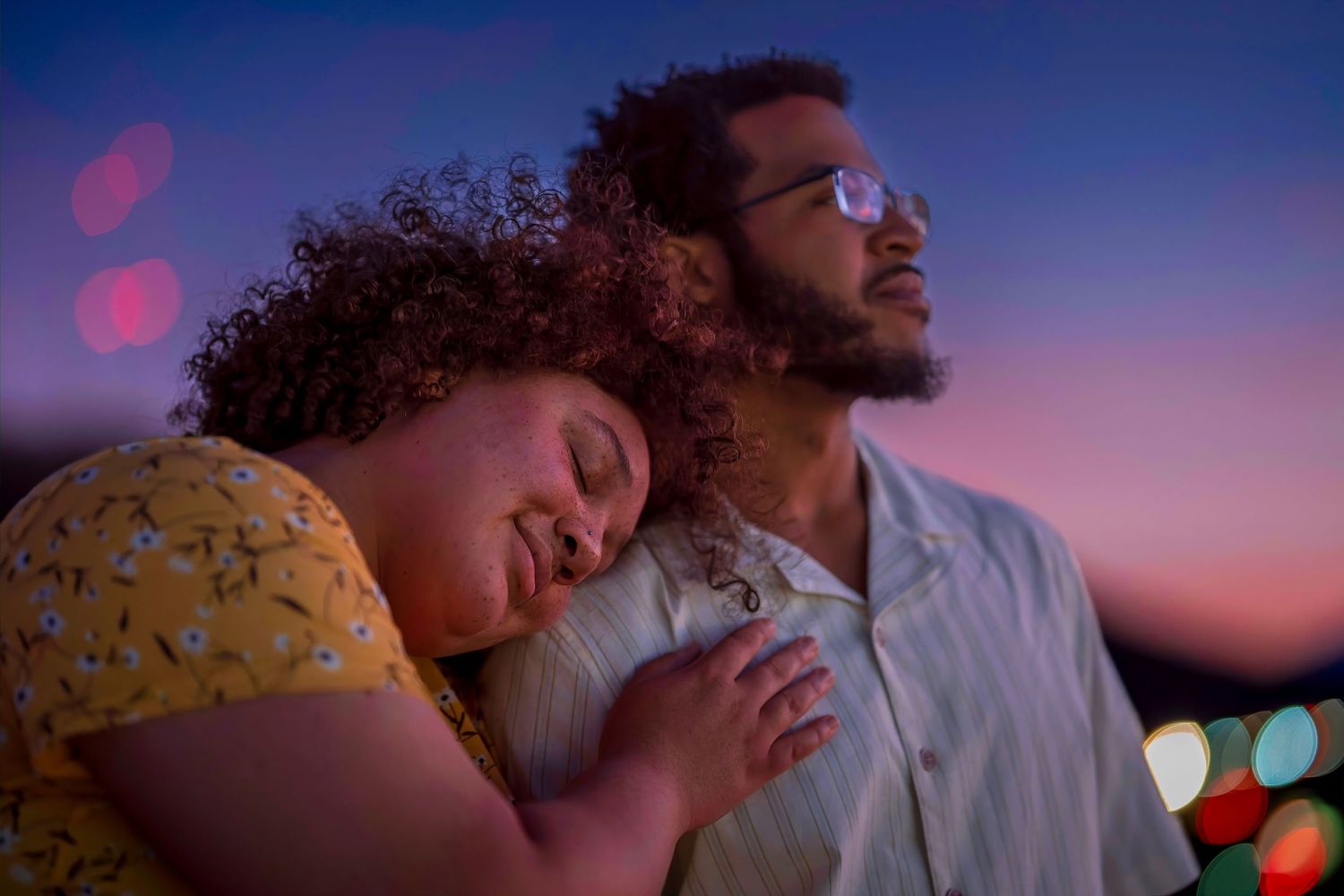 A woman rests her head on a man's shoulder at dusk, sharing a tender romantic moment under a purple twilight sky.