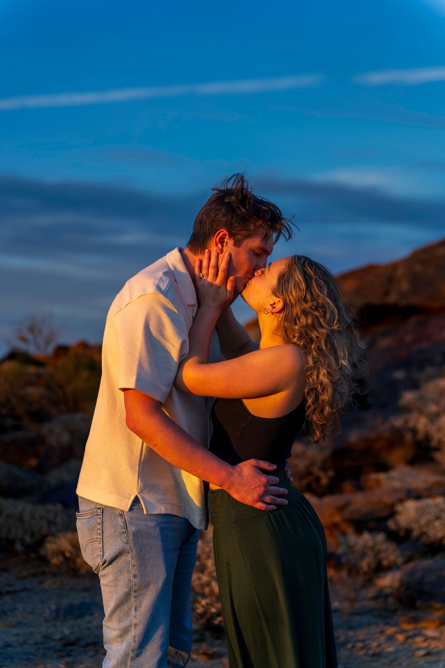 A couple shares a passionate kiss outdoors during golden hour with rocky terrain behind them.