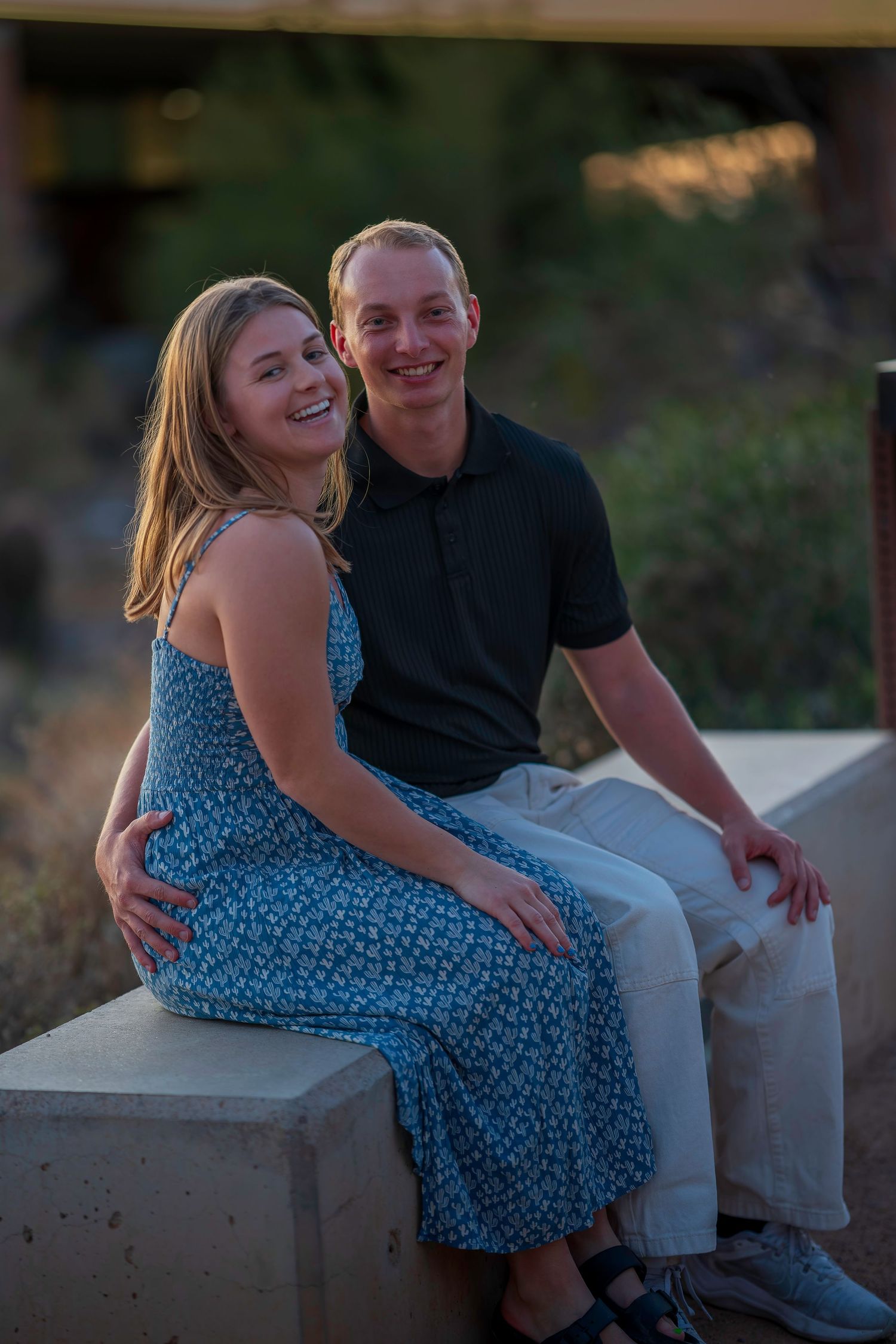 A smiling couple sits together on a concrete ledge at sunset.