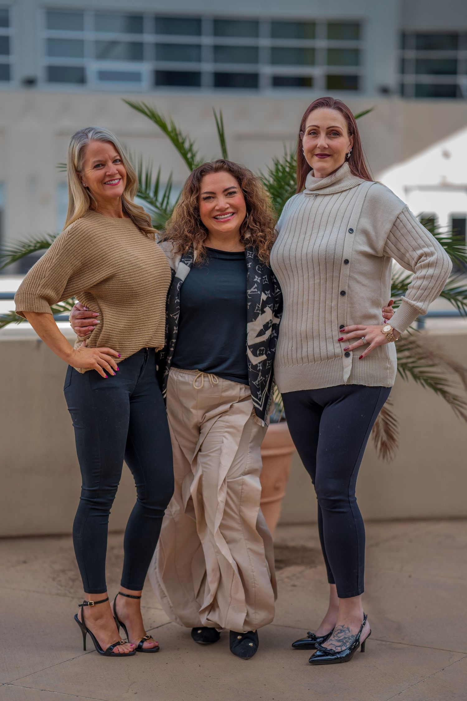 Three women in casual outfits posing together outdoors with modern building in background.