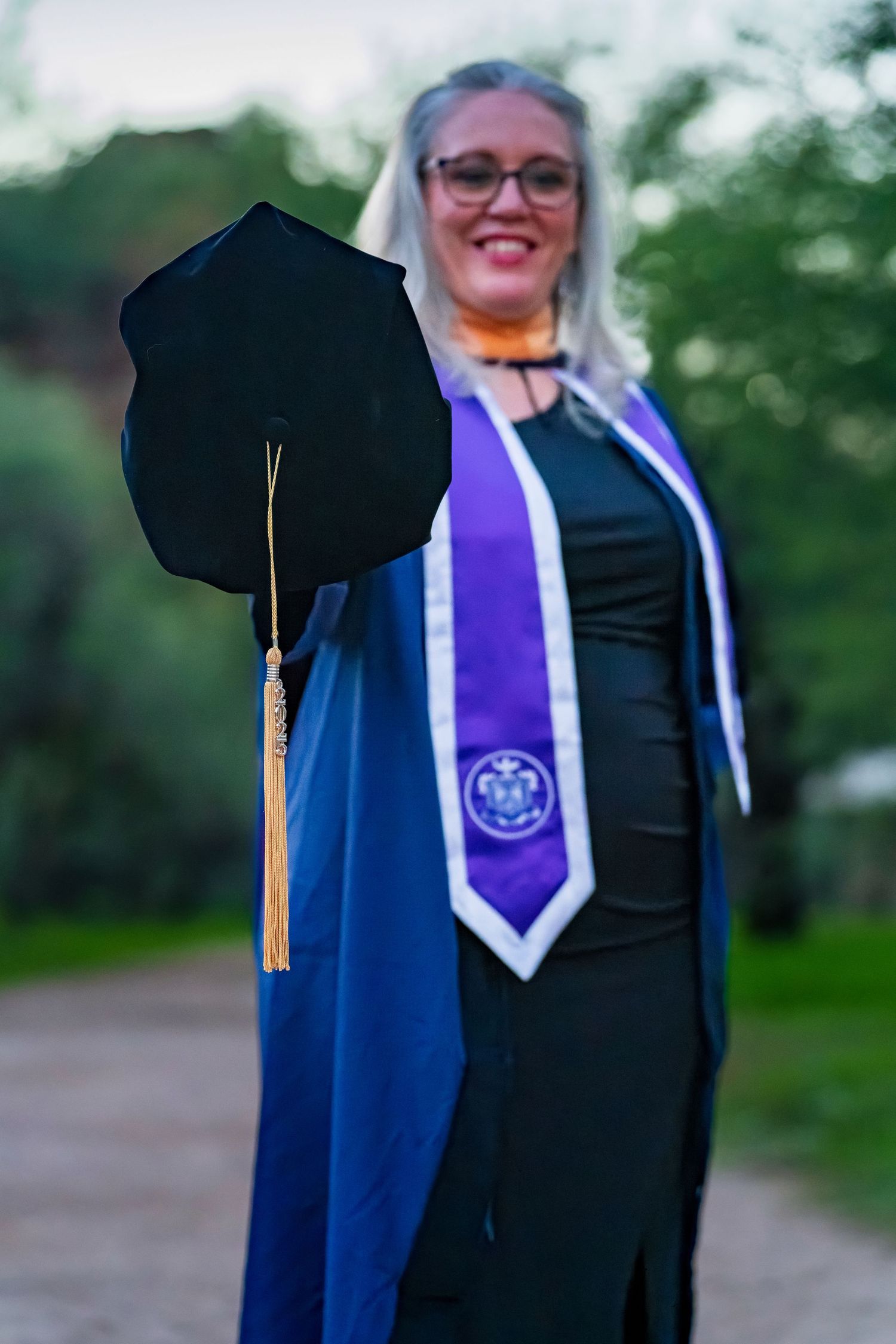 Academic regalia details showing blue gown with purple stole and gold tassel against natural backdrop.