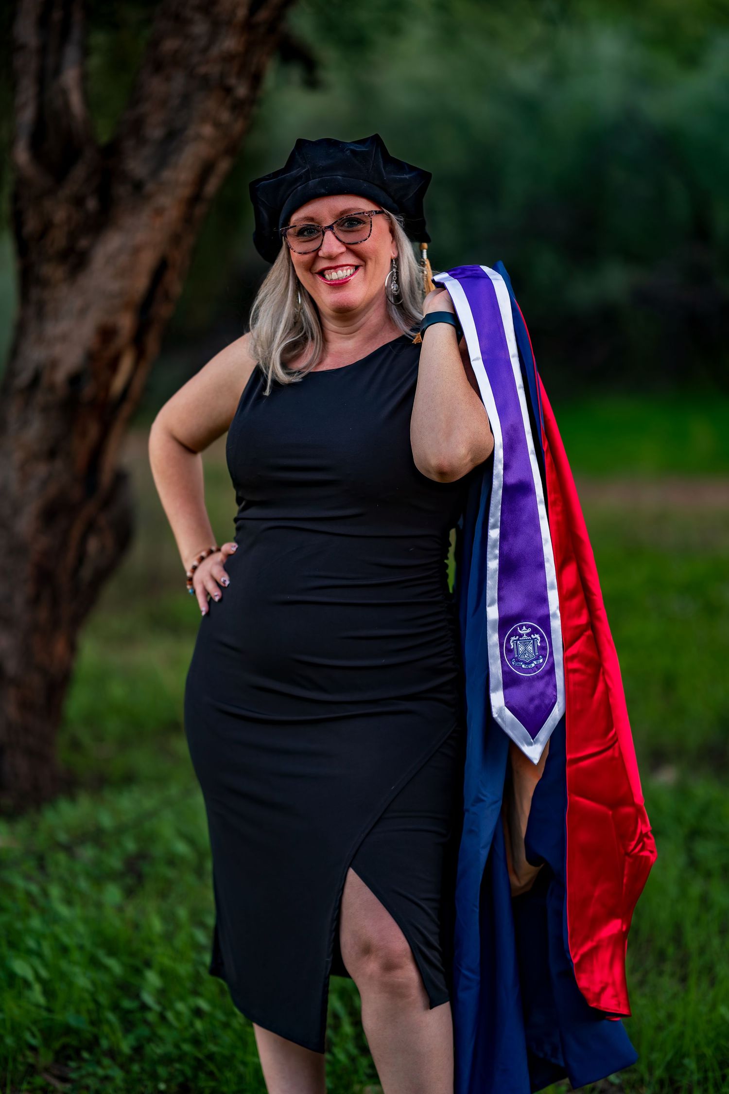 Graduate in black dress and academic cap poses with colorful graduation stoles outdoors.