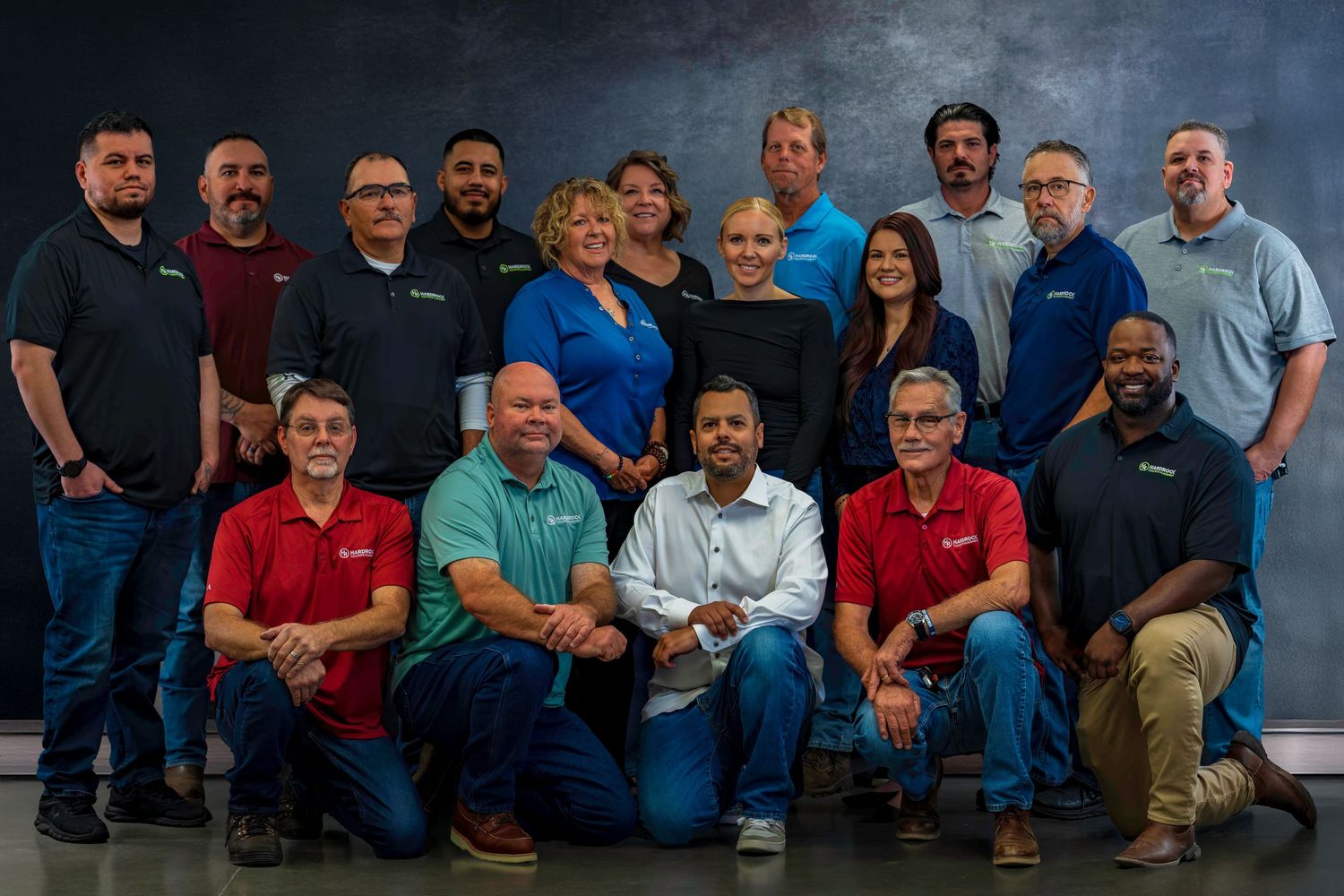 Large team photo of fifteen employees arranged in two rows against dark studio background.