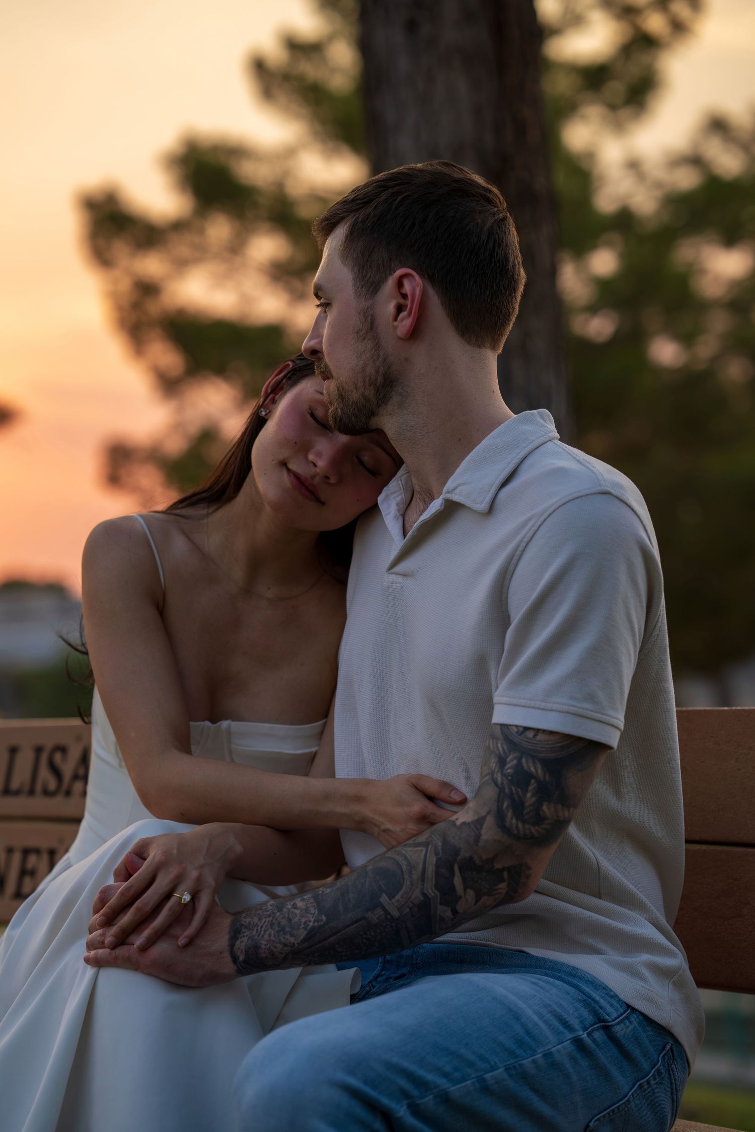 Couple shares tender moment on bench at sunset, man with tattoos embracing woman in white dress.