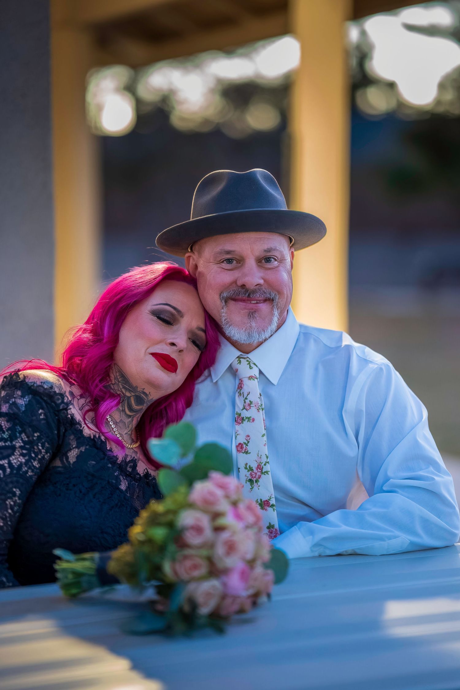 Couple posing together at a table with pink roses bouquet, woman with pink hair leaning on man in gray hat and floral tie.