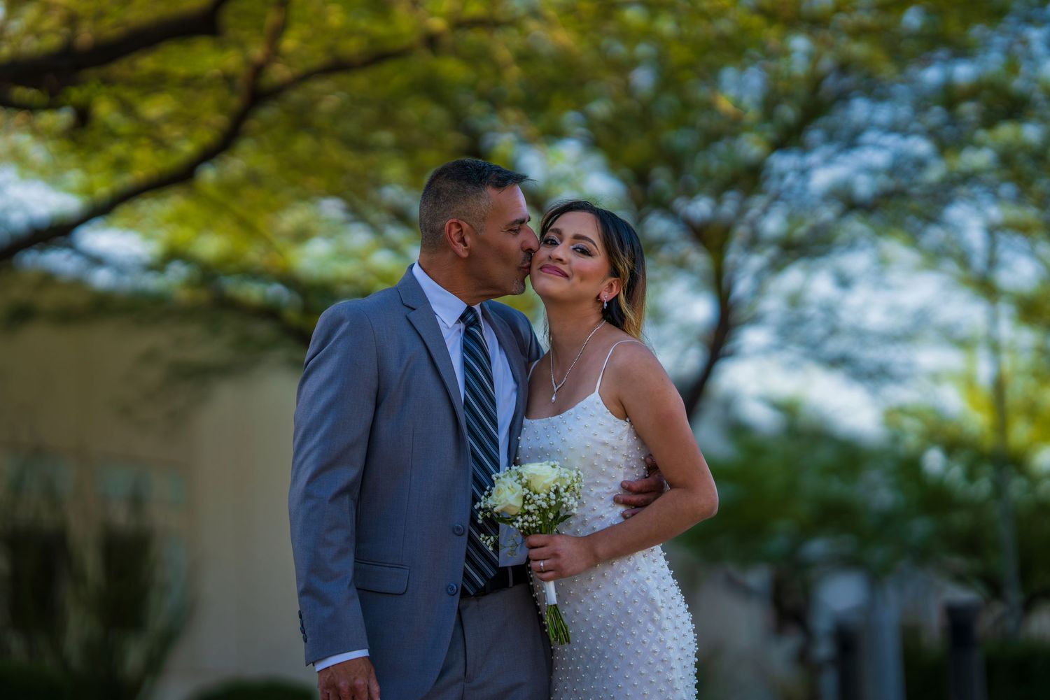 A groom in a gray suit kisses his bride on the cheek as she holds a bouquet outdoors.