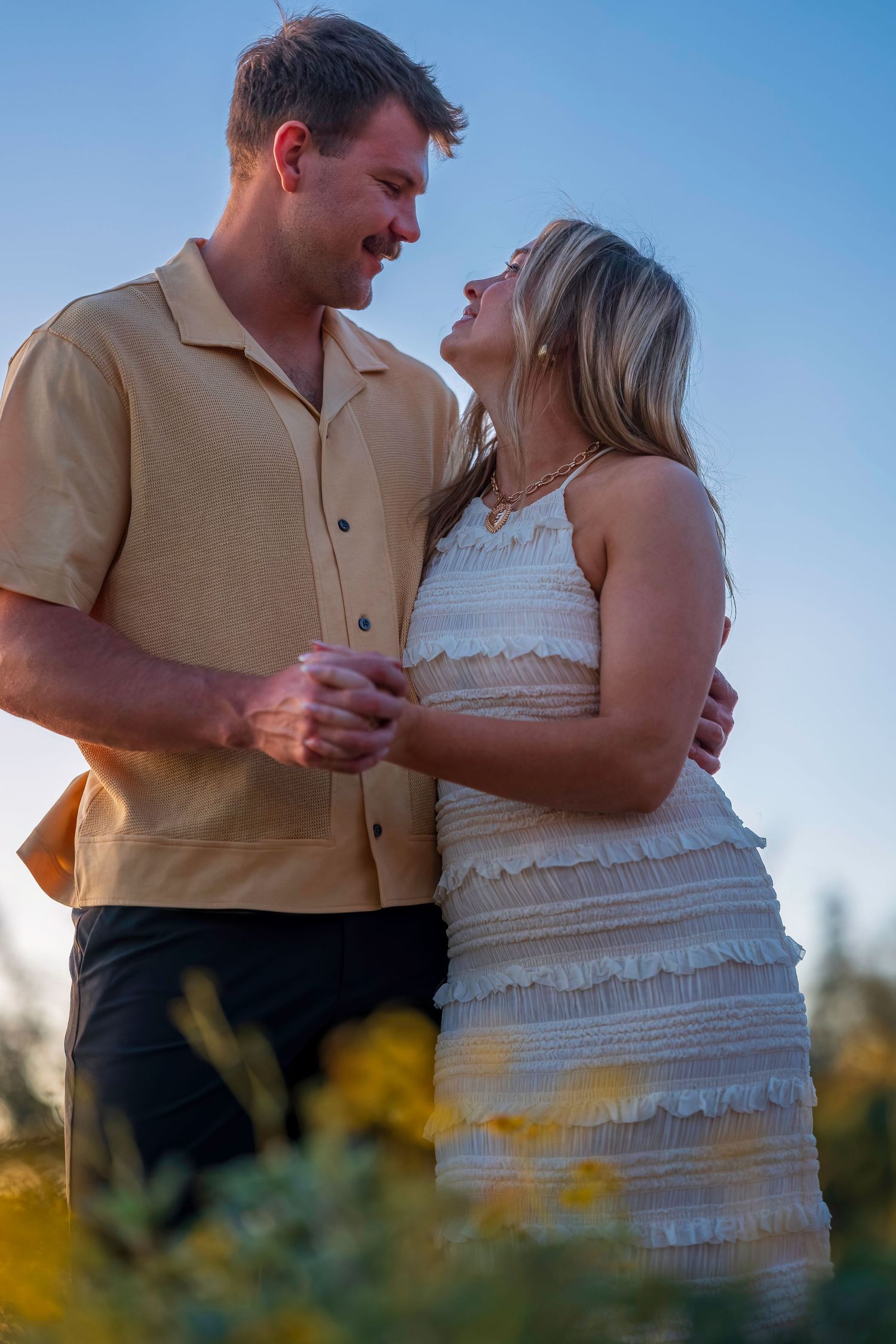 A couple gazes lovingly at each other outdoors, man in beige shirt, woman in white ruffled dress, yellow flowers foreground.