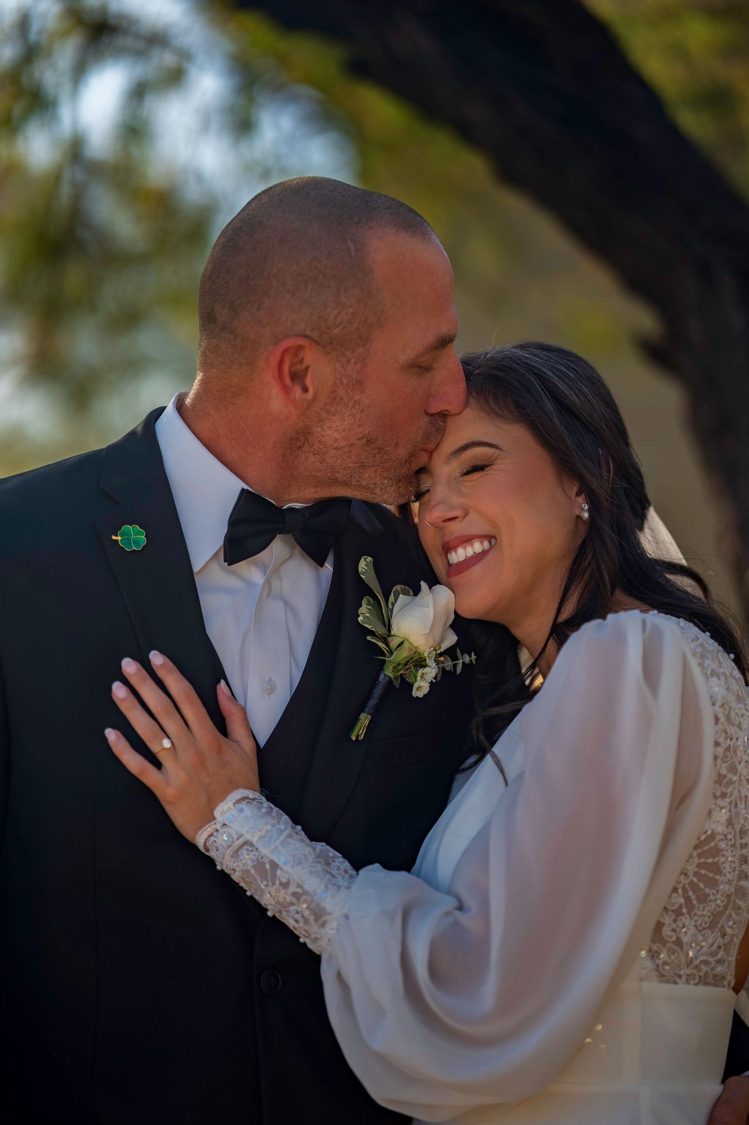 Groom in black tuxedo kisses smiling bride in white long-sleeve wedding dress outdoors.