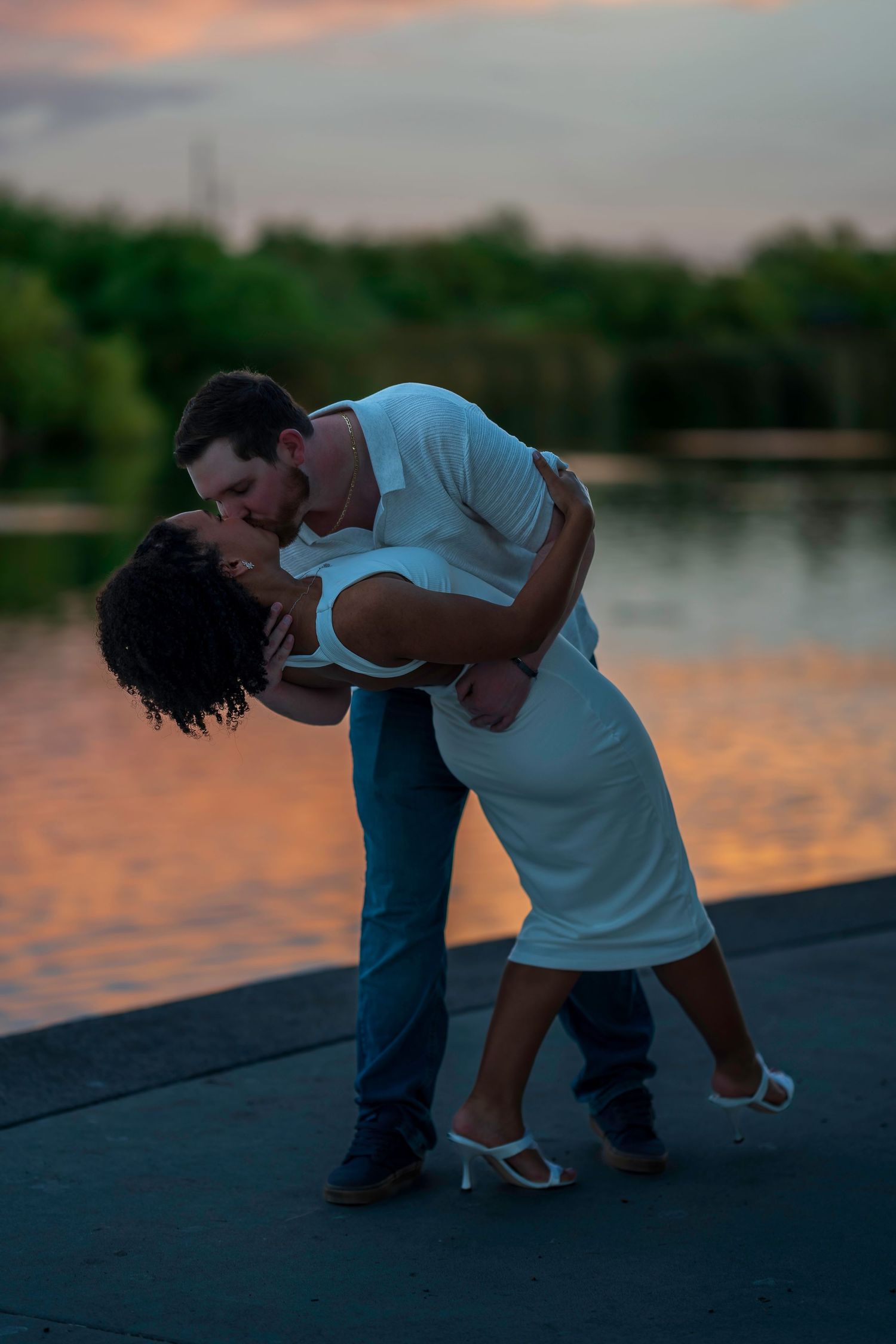 Couple sharing a romantic dip kiss by a lake at sunset, man in white shirt and jeans, woman in white dress.