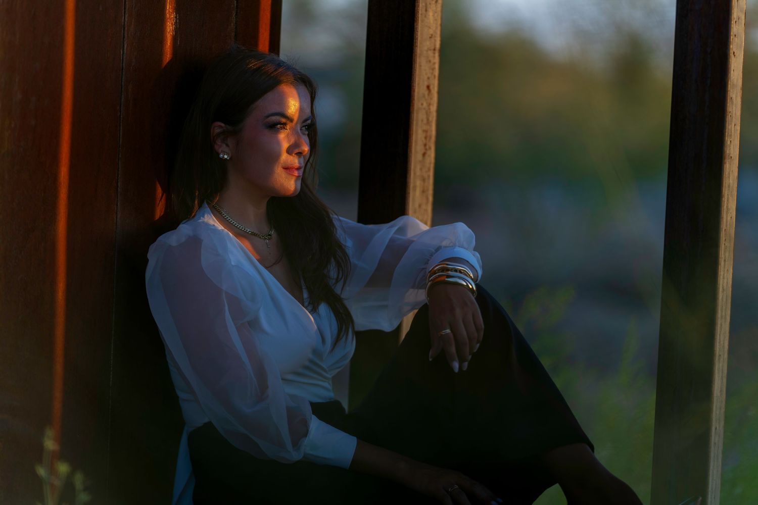 A young woman in a white blouse sits pensively against wooden beams in warm golden hour light.
