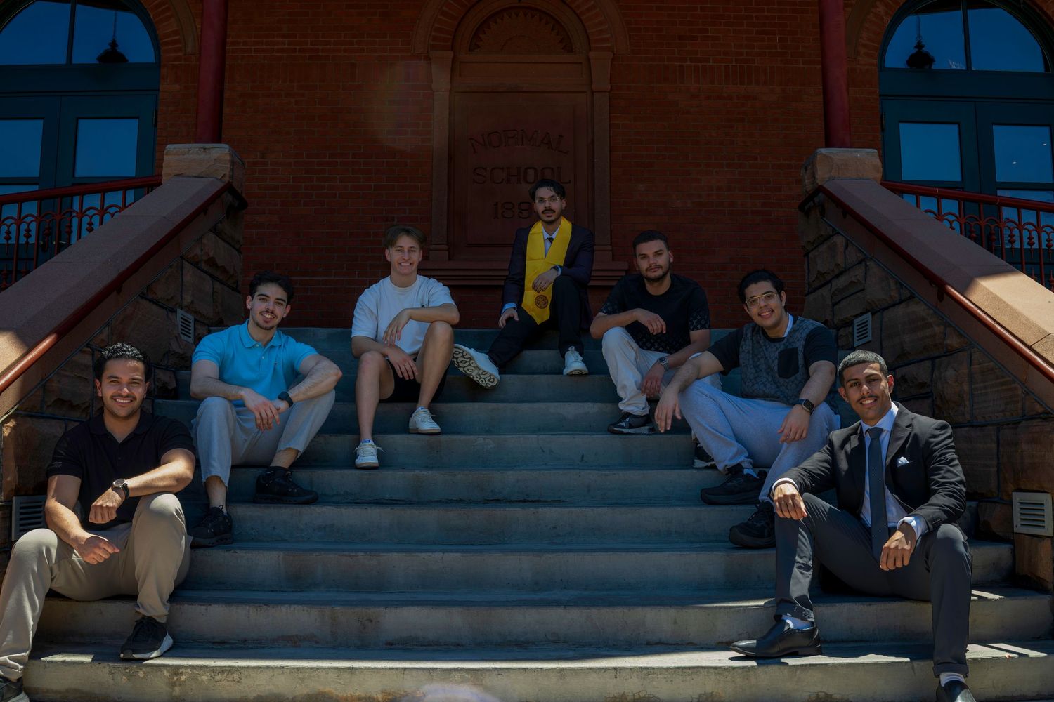Group of seven young adults sitting on brick building steps, posing for a casual outdoor photo in sunny weather.
