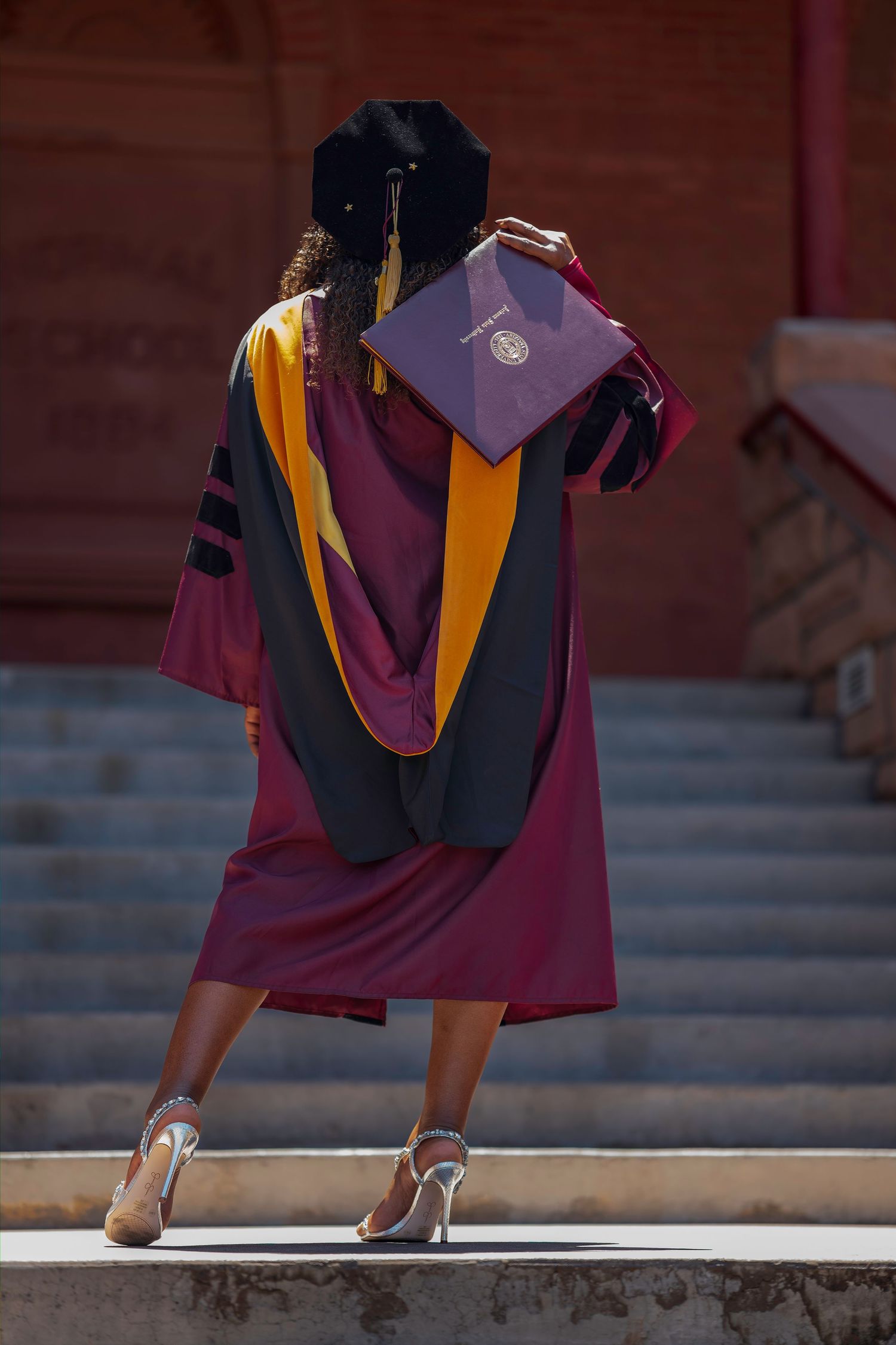 Graduate in maroon and black doctoral regalia holds diploma while standing on steps, viewed from behind.