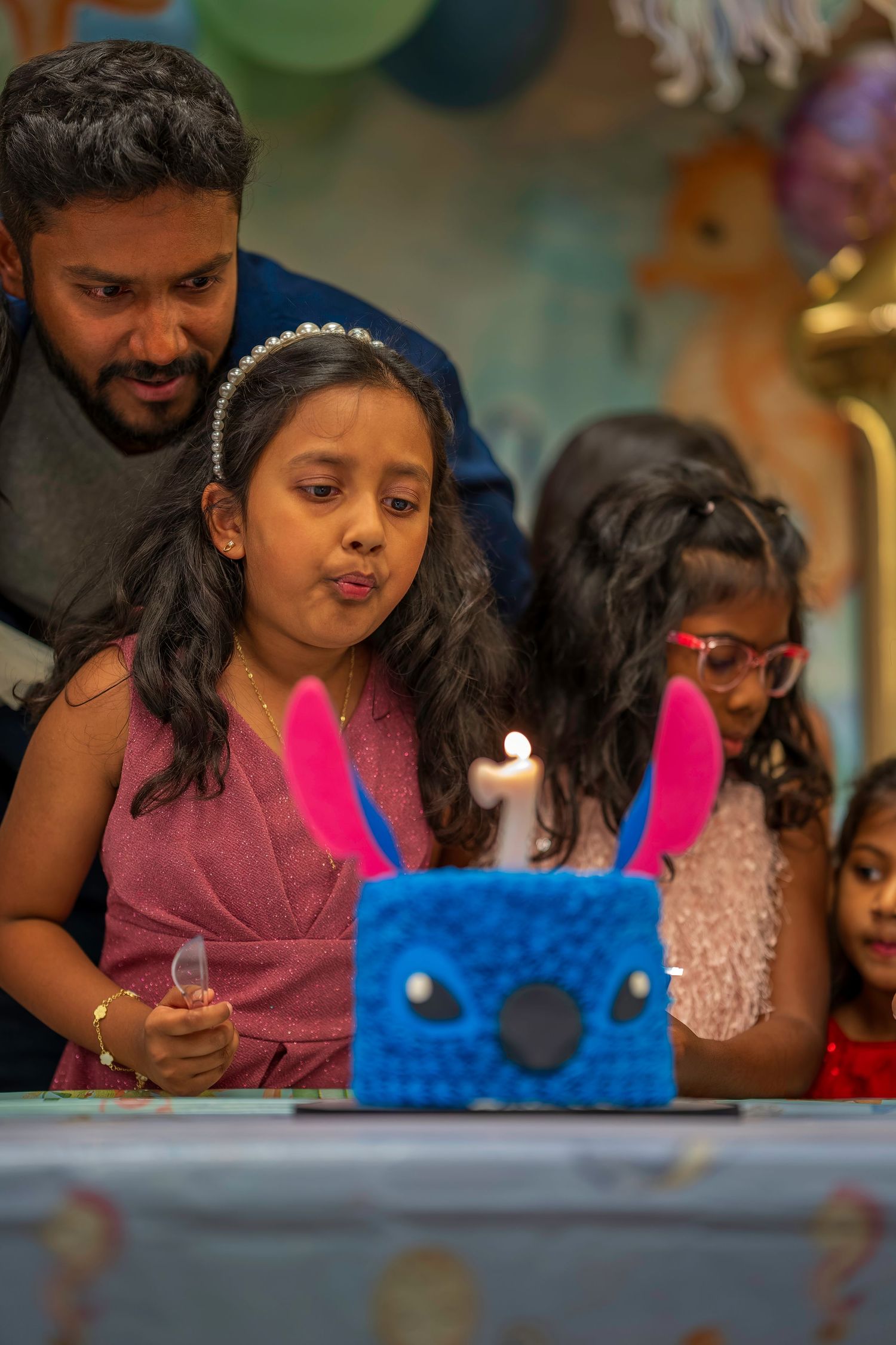 A young girl blows out a candle on a blue bunny birthday cake while a man looks on at a birthday party.