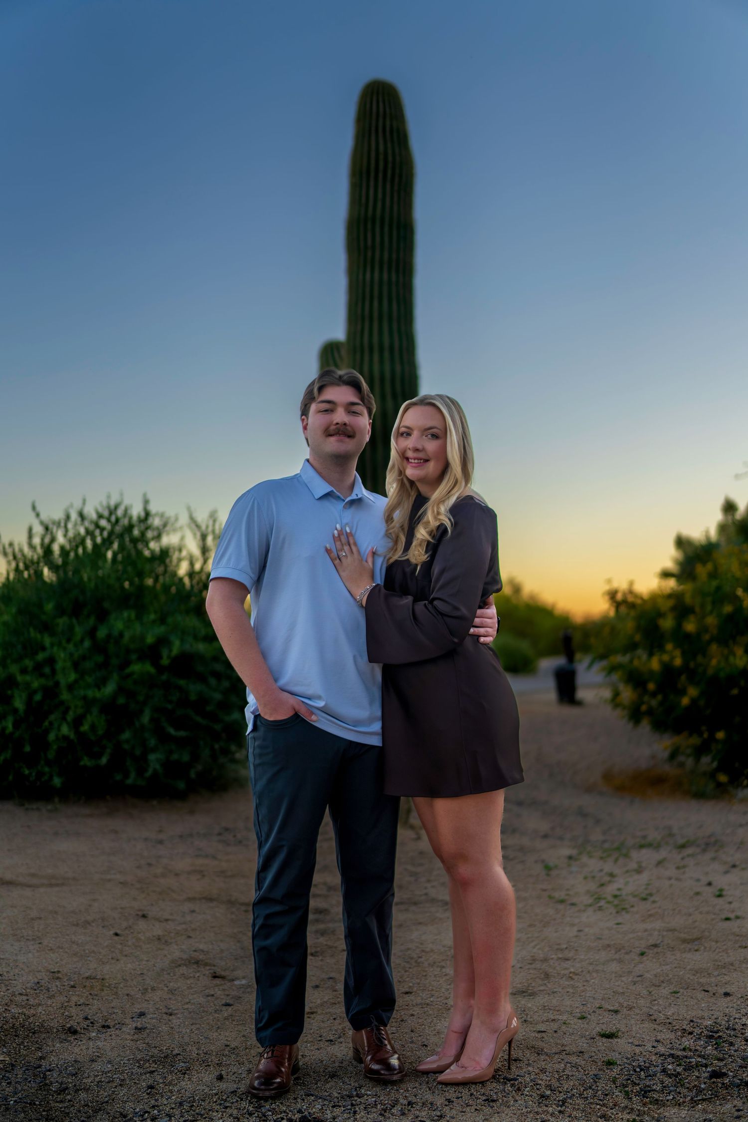 An engaged couple poses at sunset in the Arizona desert, with a tall saguaro cactus behind them, as the woman shows her ring.