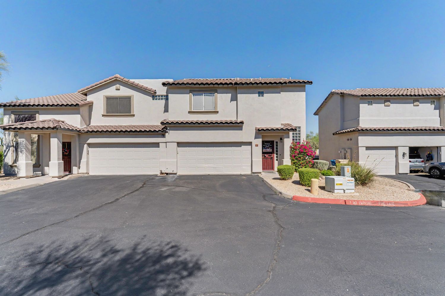 Two-story beige stucco home with attached two-car garage and red accents in a sunny Arizona neighborhood.