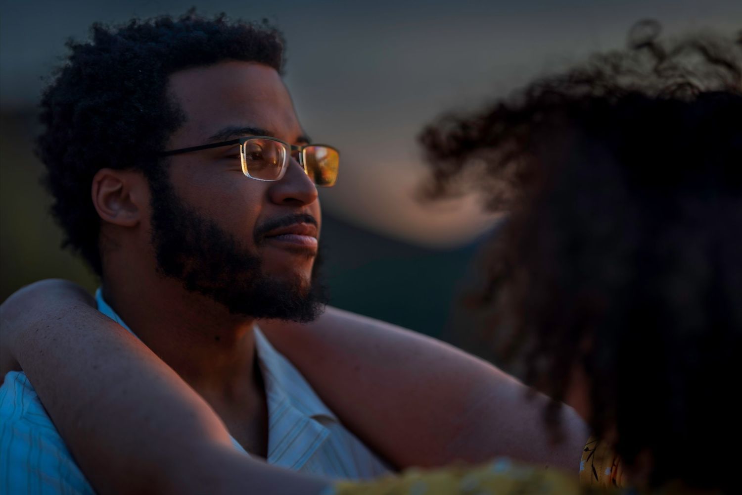 A young Black man with glasses and a beard gazes thoughtfully at dusk, with his arm around someone.