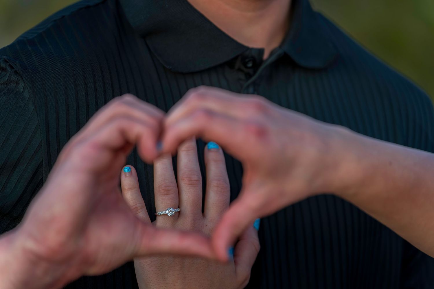 Close-up of couple forming heart shape with hands displaying diamond engagement ring with blue nails.