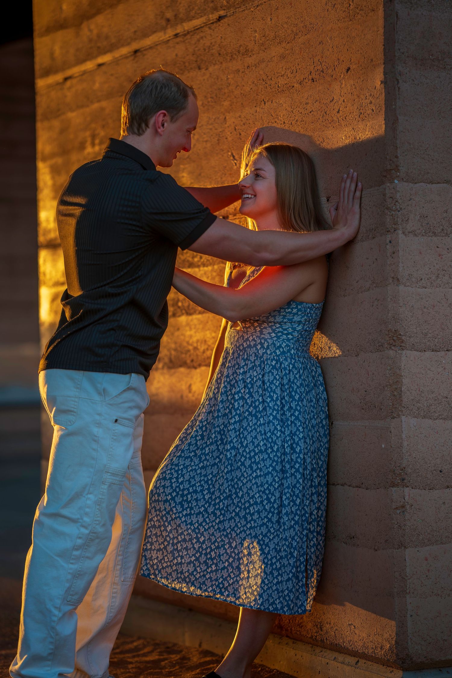 A couple shares a romantic moment leaning against a stone wall at sunset.
