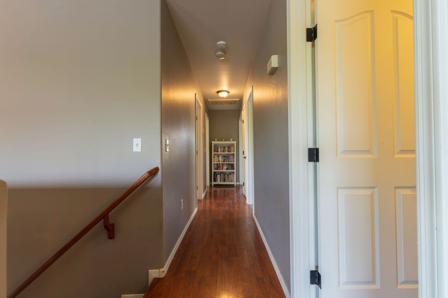 Bright hallway with hardwood floors, gray walls, white paneled door, and wooden staircase railing leading to a bookshelf.