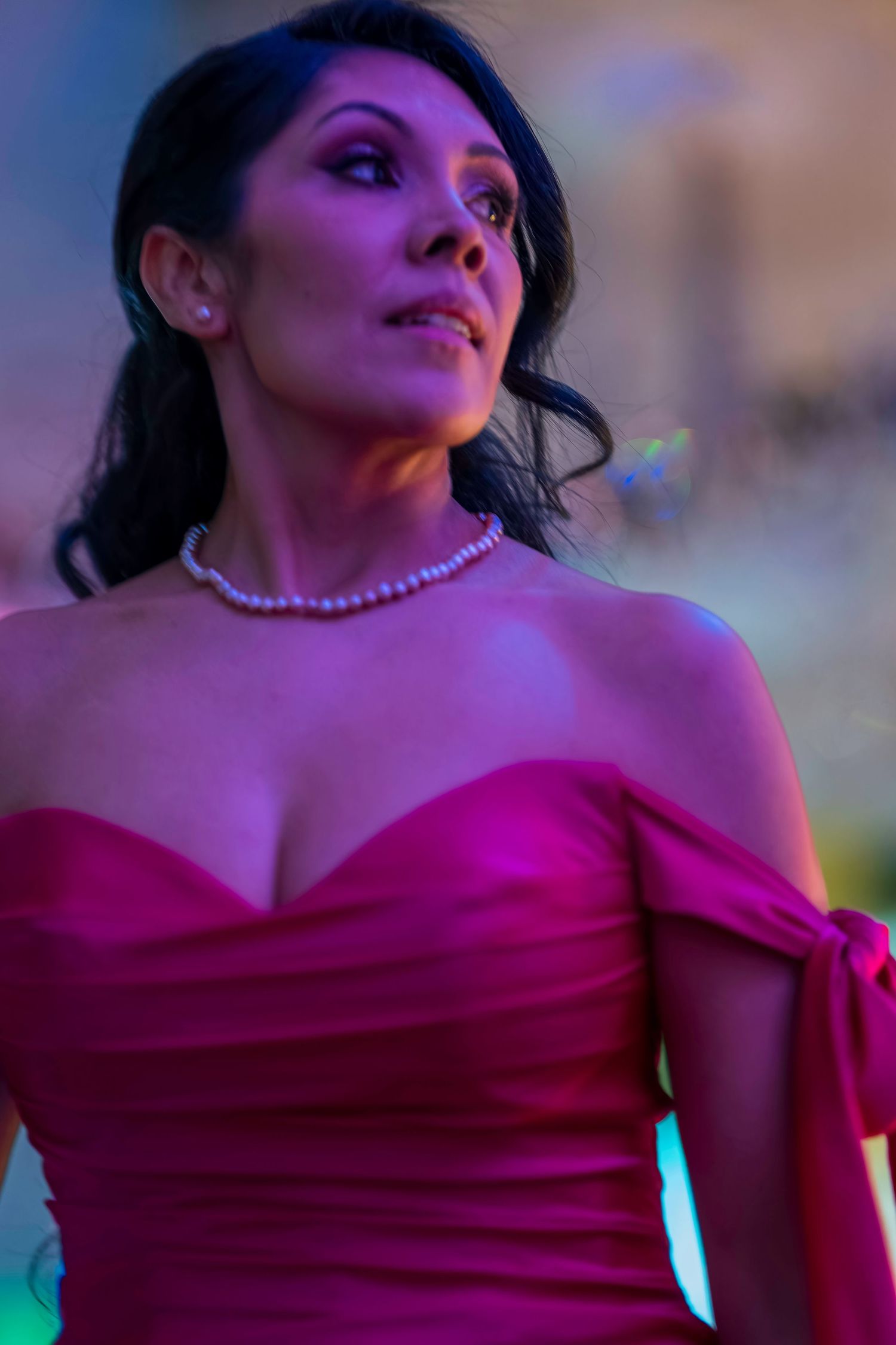 A woman in a red off-shoulder gown with pearl necklace poses elegantly under colorful lighting.