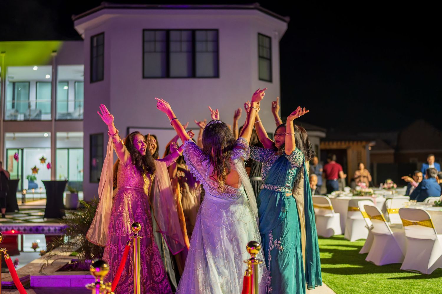 People in colorful evening gowns dance with raised arms at an outdoor party venue.